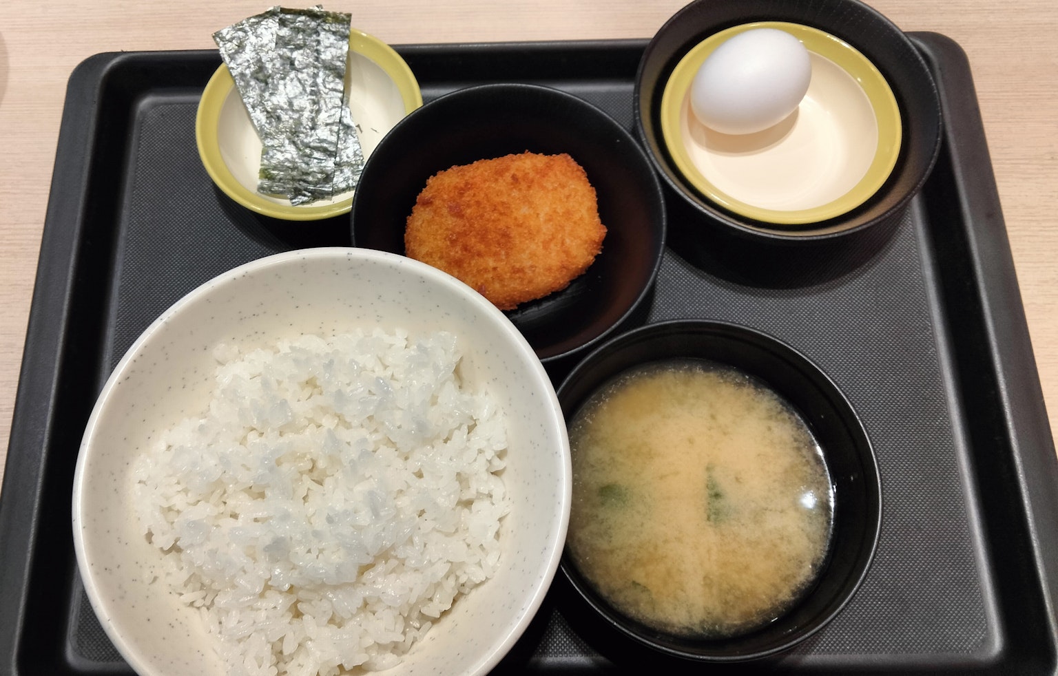 Una bandeja de comida japonesa con arroz, sopa de miso, un huevo, una croqueta y un artículo envuelto en papel de aluminio