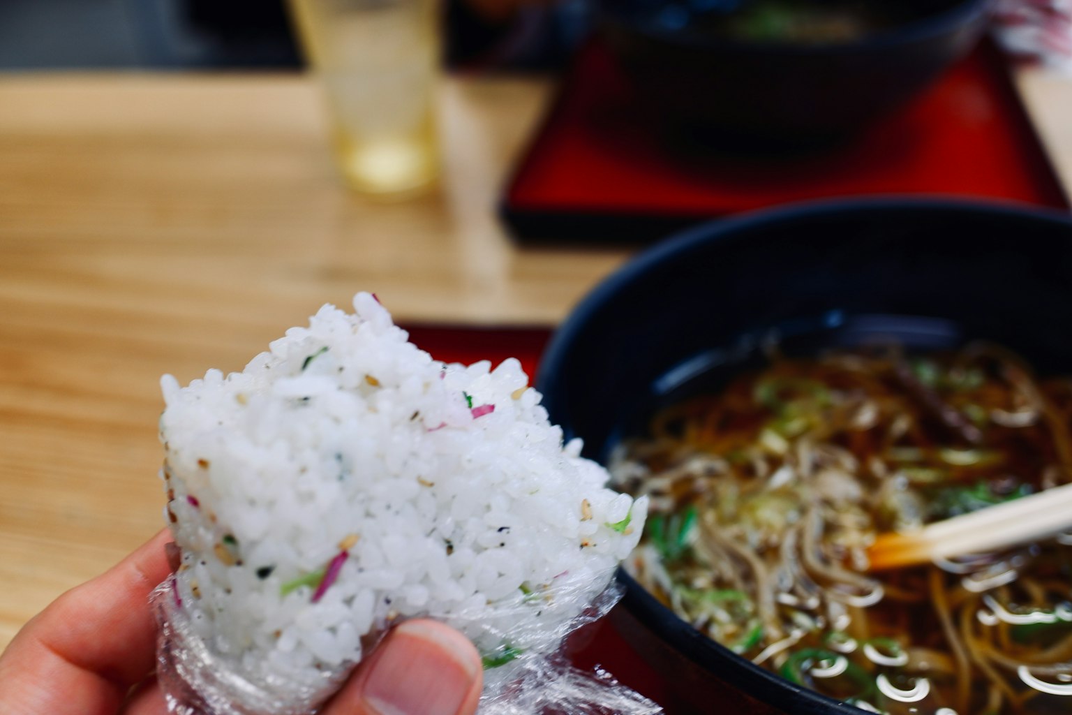 Eine Hand hält ein Onigiri mit einer Schüssel Nudeln im Hintergrund