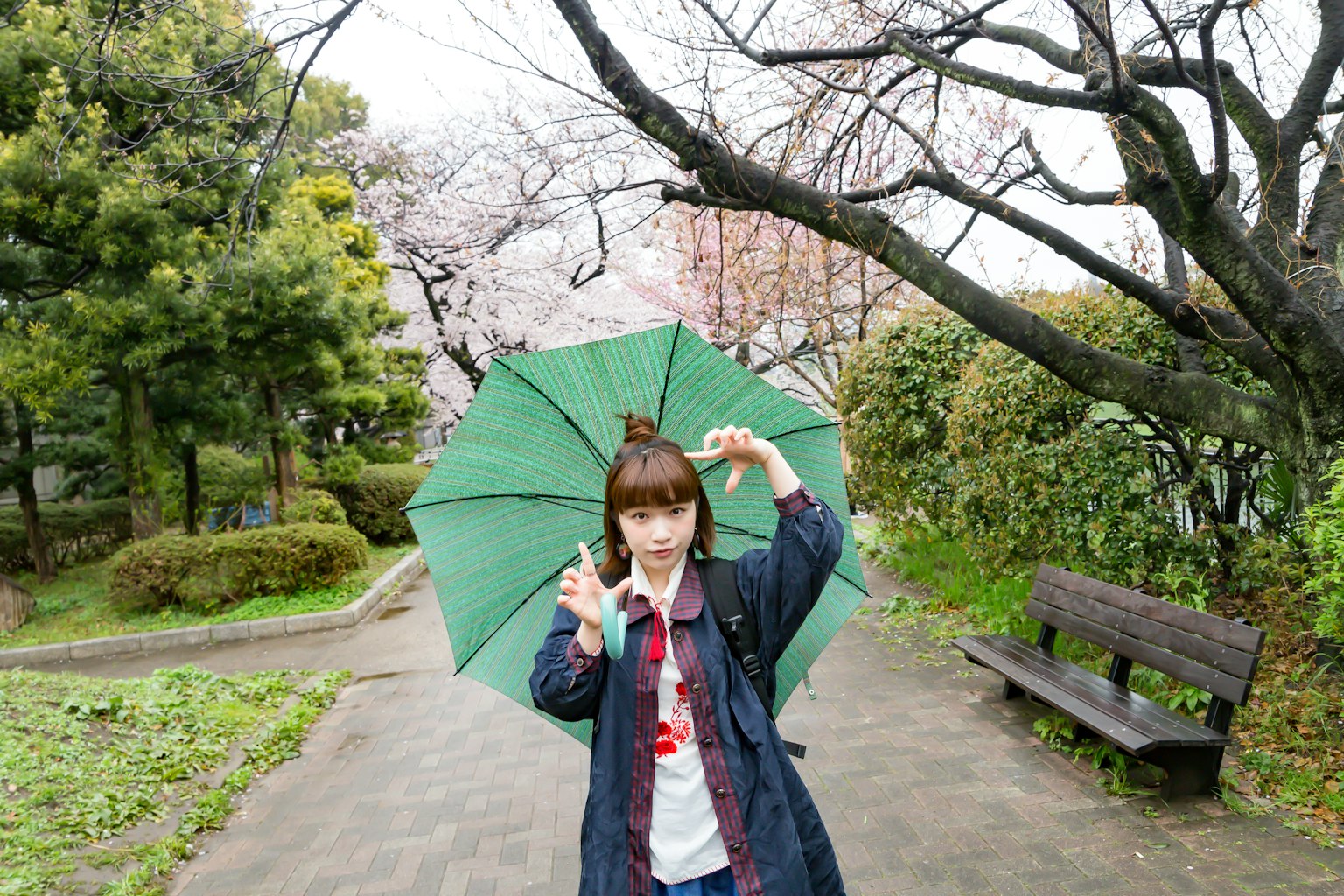 Girl holding a green umbrella in a park with cherry blossom trees and a bench