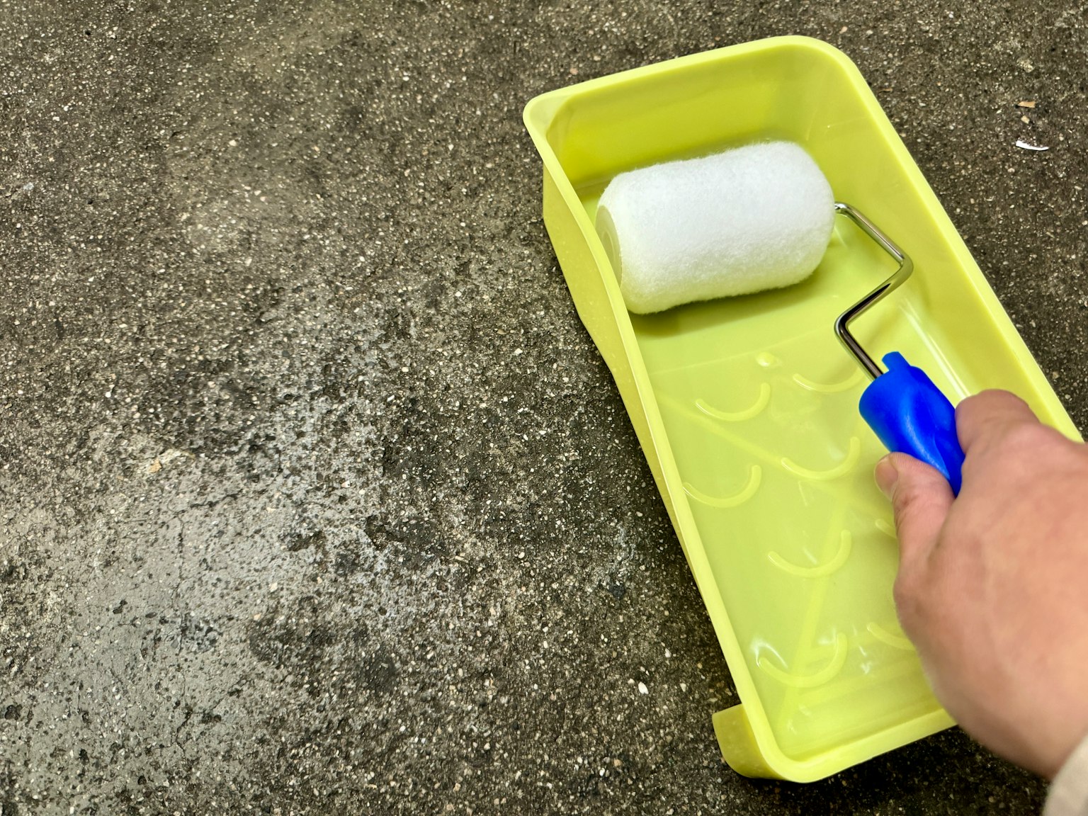 Sebuah tangan memegang roller cat dengan pegangan biru di atas baki kuning dengan roller cat putih