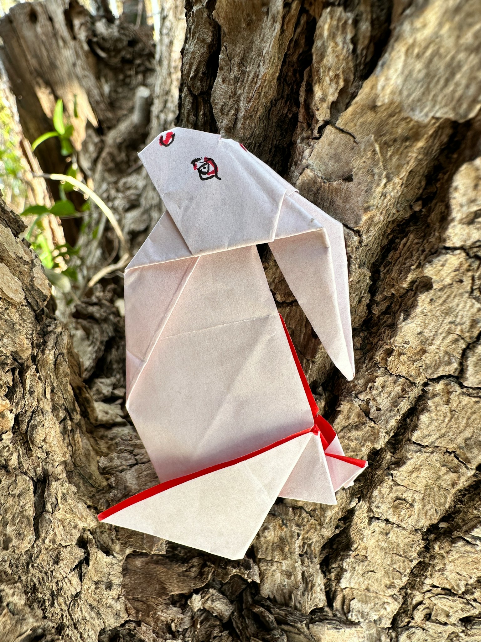 White origami rabbit with red accents attached to a tree trunk