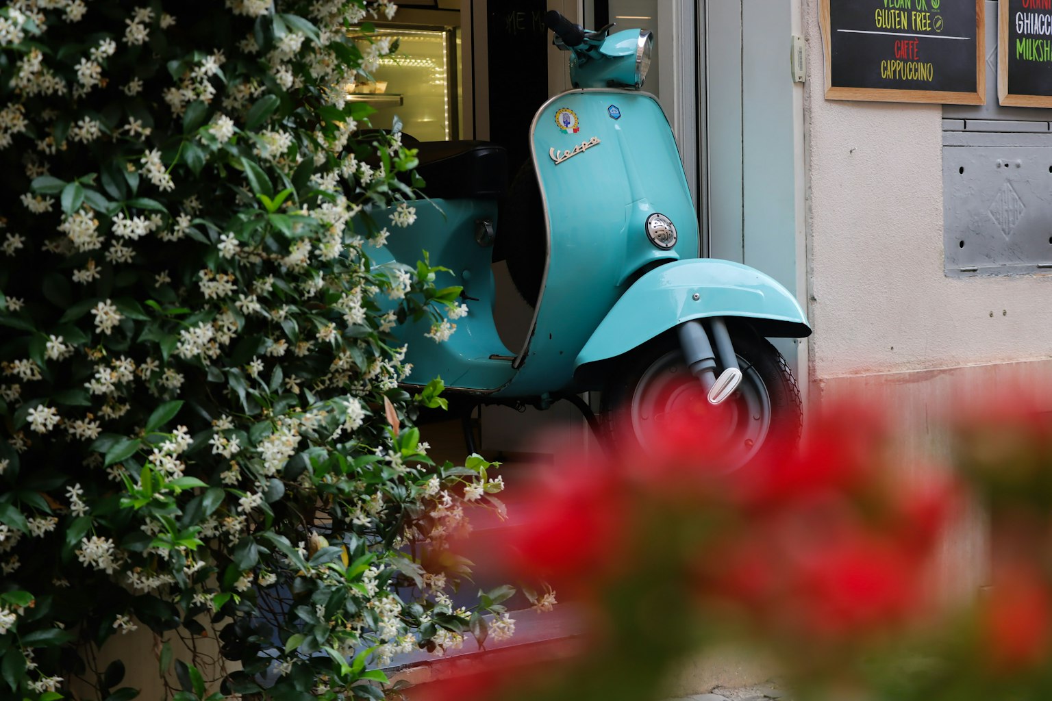 Ein blauer Roller teilweise hinter Blumen versteckt mit einem Café-Eingang im Hintergrund