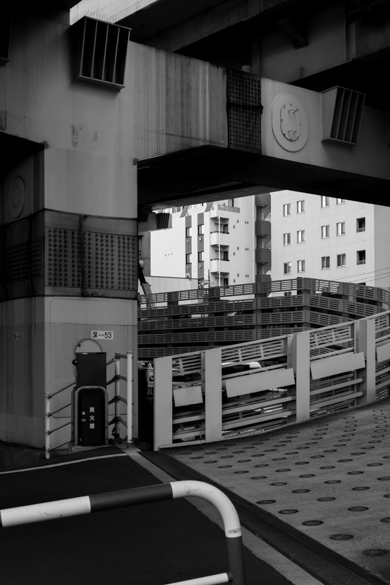 Scena urbana in bianco e nero con una struttura di parcheggio sotto un cavalcavia edifici visibili sullo sfondo