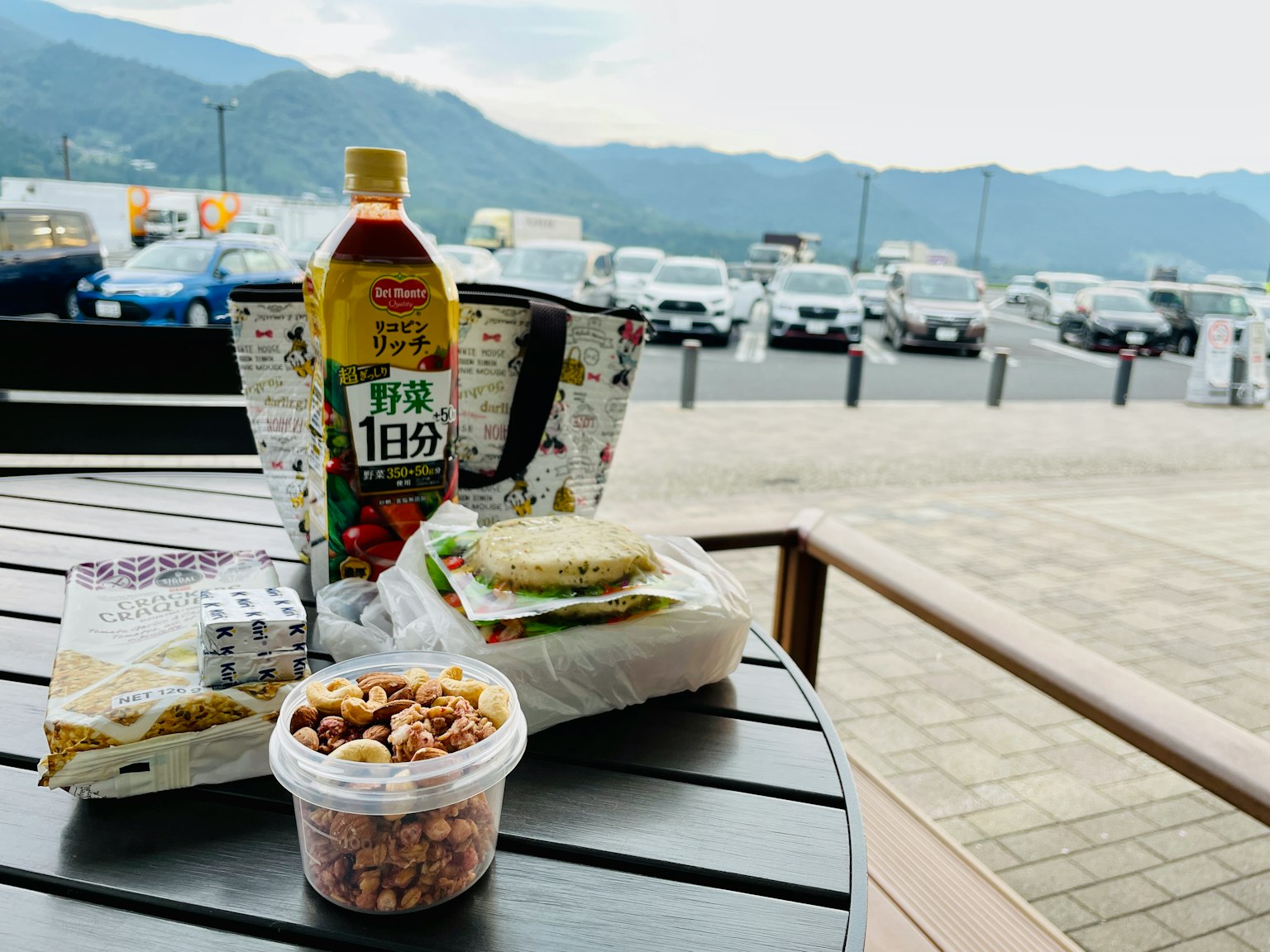 Sebuah meja dengan sandwich, camilan, dan minuman dengan latar belakang pegunungan dan tempat parkir