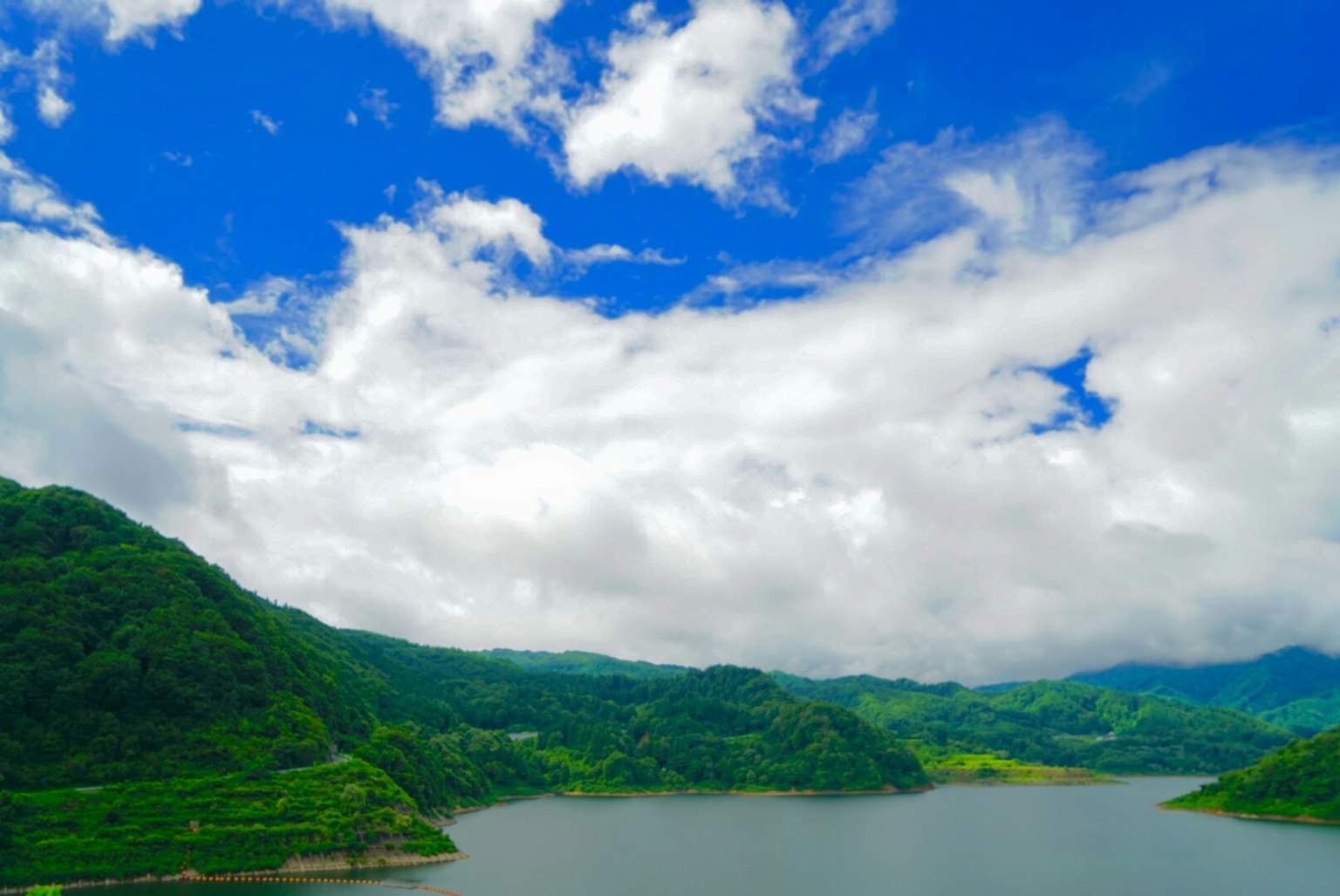Scenic view of a lake surrounded by green mountains under a blue sky with white clouds