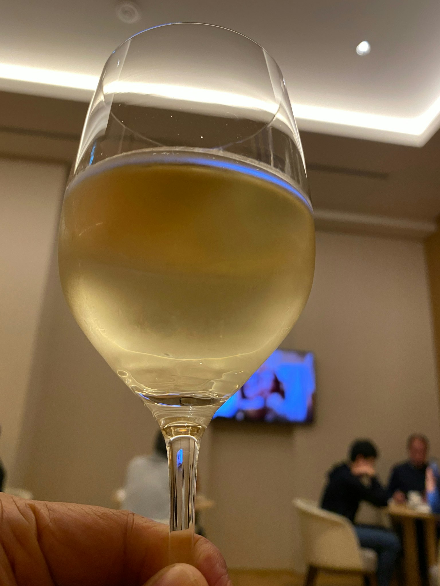 Una mano che tiene un bicchiere di vino bianco con persone e una TV sullo sfondo