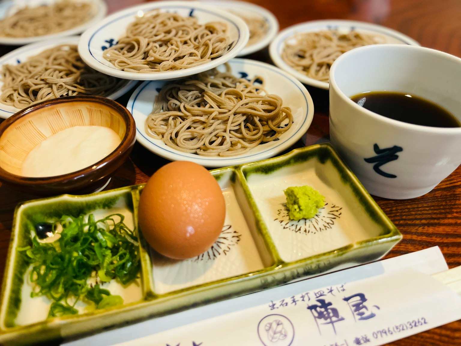 Japanese meal featuring soba noodles, a bowl of dipping sauce, and condiments including egg and wasabi