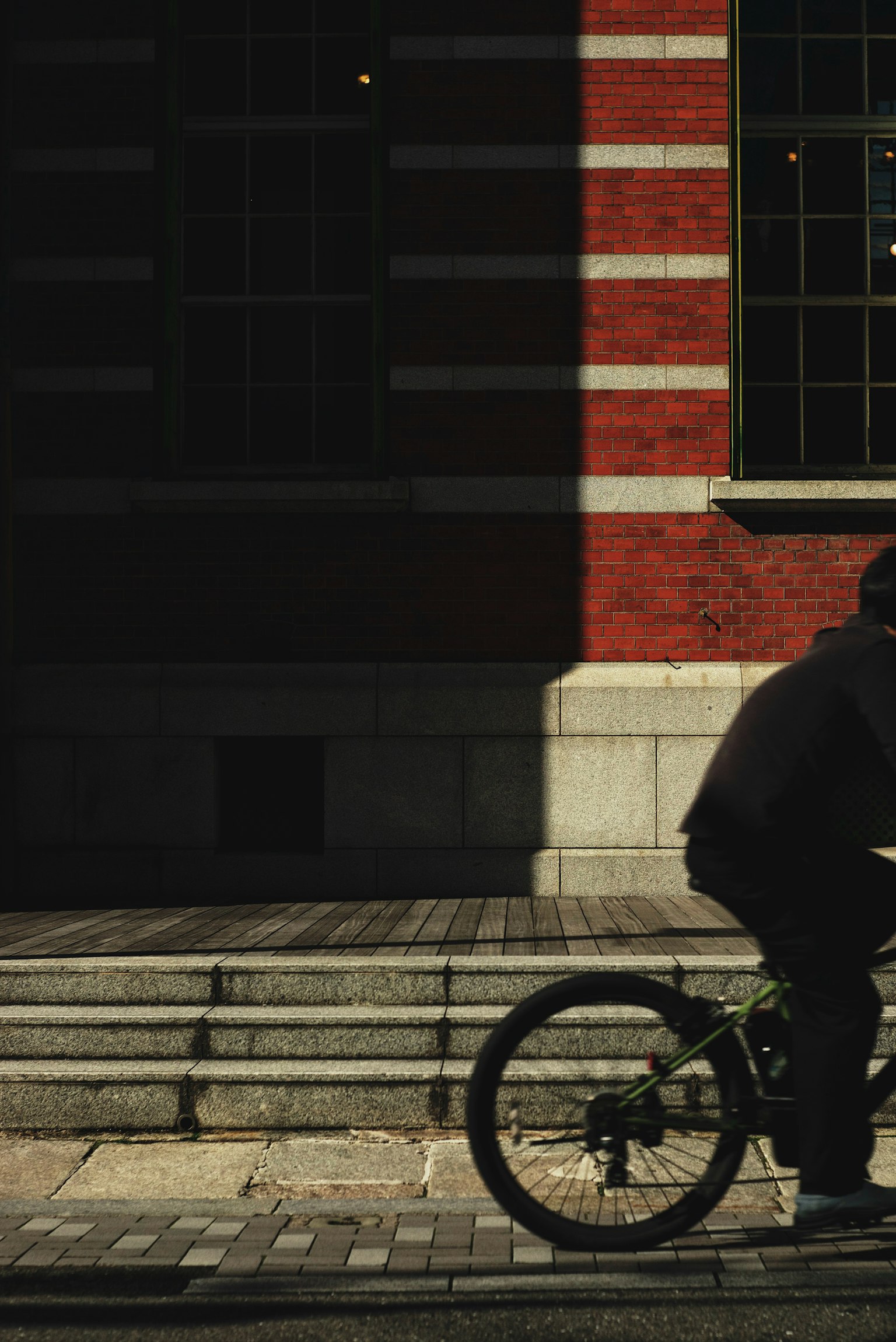 A person riding a bicycle passing in front of a red brick building in shadow