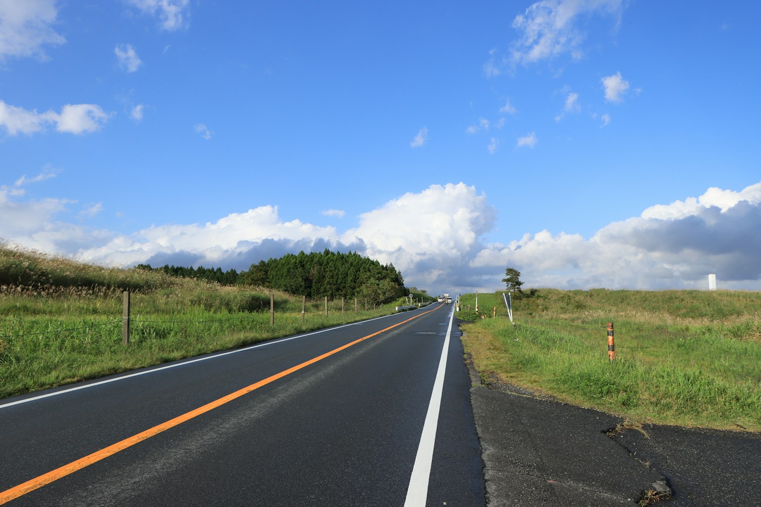 Jalan lurus di bawah langit biru dengan awan putih dan rumput hijau