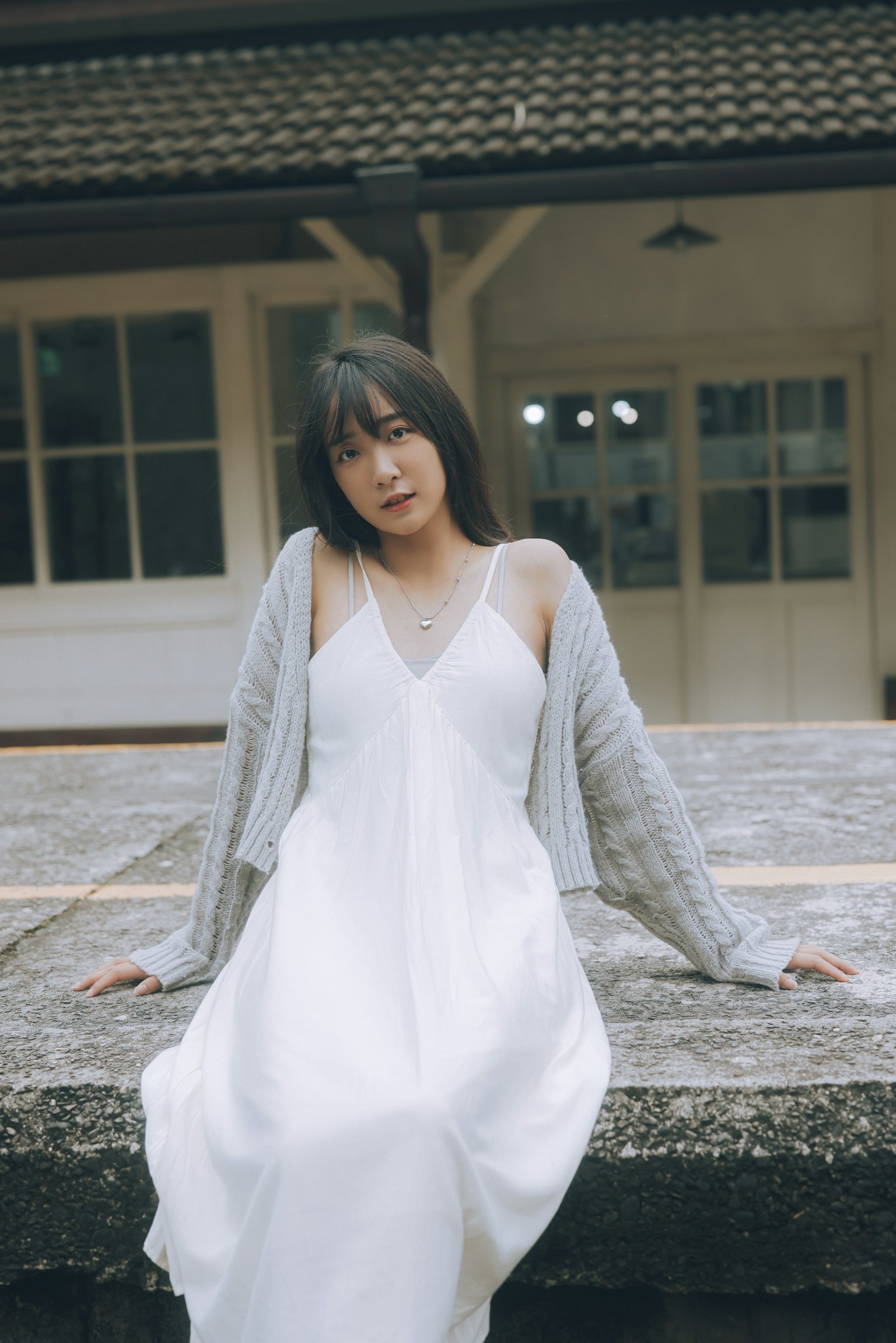 白いドレスを着た女性がグレーのカーディガンを羽織り、建物の前でポーズをとっている