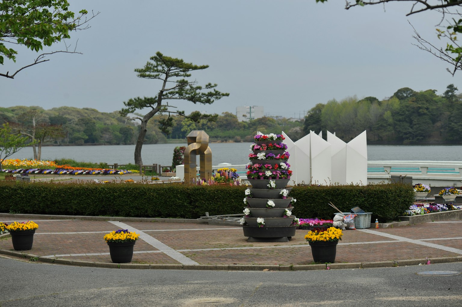 Scena di parco con vasi di fiori disposti lungo i sentieri e un lago con sculture bianche sullo sfondo