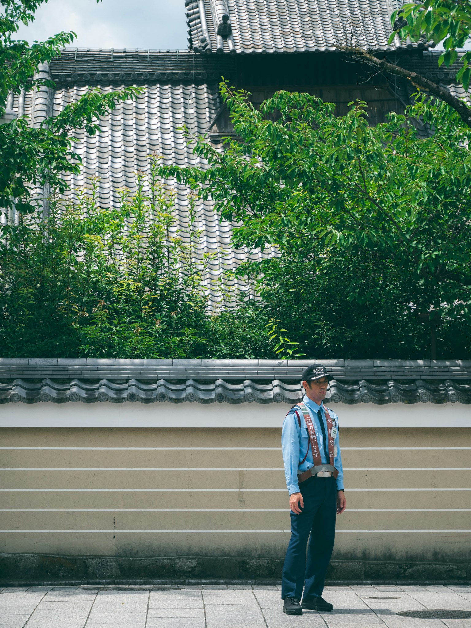 A security guard in a blue uniform standing in front of a wall with green trees in the background