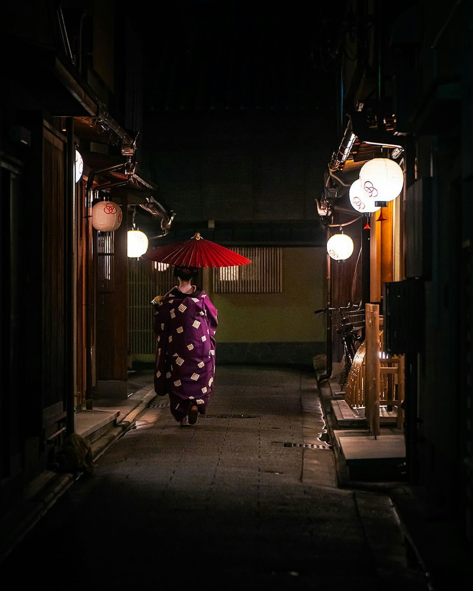 Một người phụ nữ trong trang phục kimono đi bộ vào ban đêm với chiếc ô đỏ