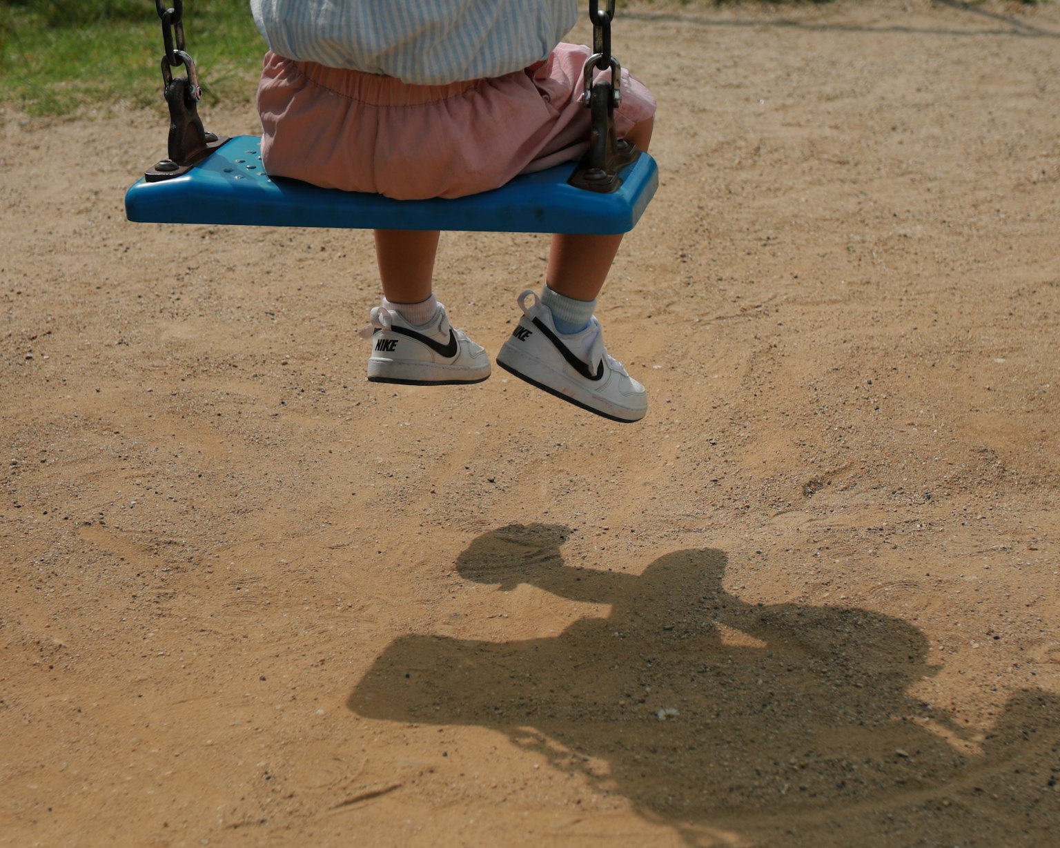 เท้าของเด็กที่แกว่งอยู่บนชิงช้าสีฟ้ากับเงาบนสนามเด็กเล่นที่เป็นทราย