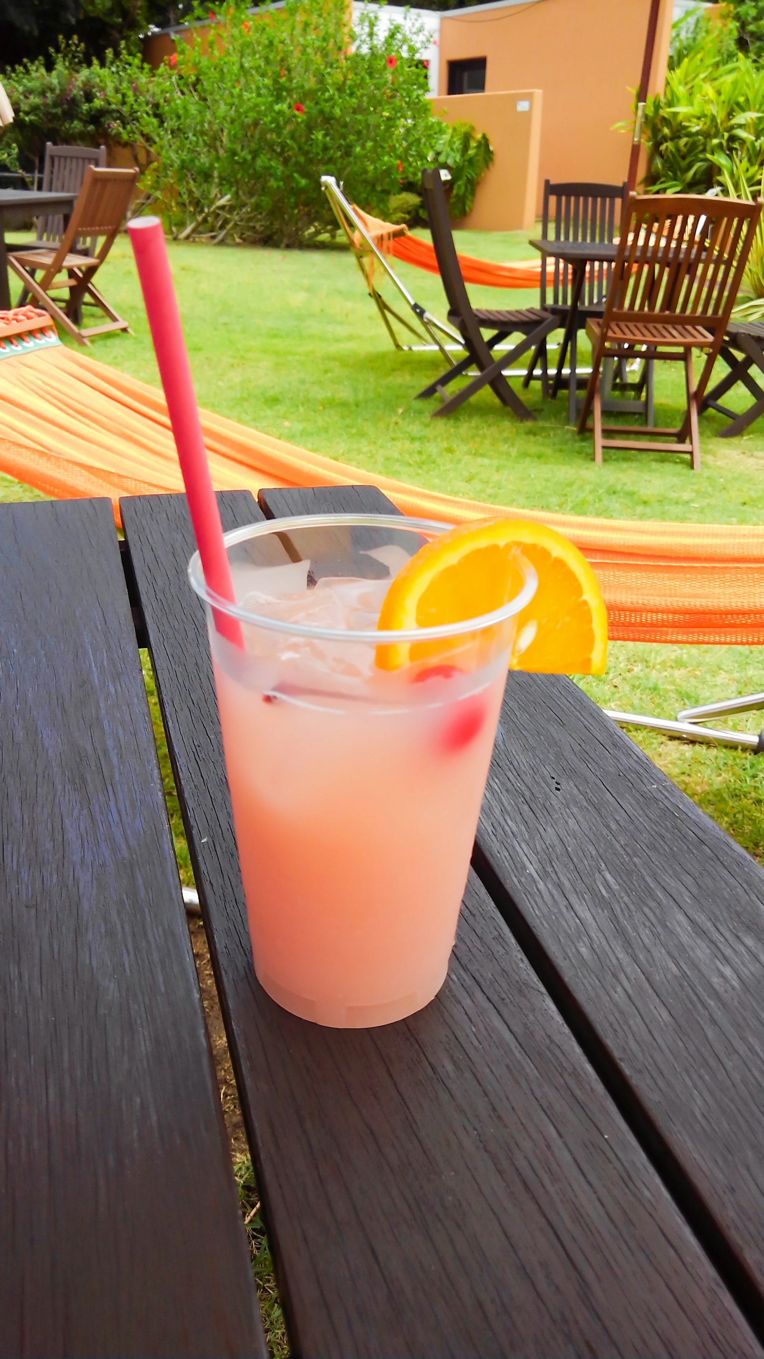 Ein Cocktail in einem Plastikbecher mit einer Orangenscheibe und einem roten Strohhalm auf einem Holztisch mit grünem Gras und Hängematten im Hintergrund