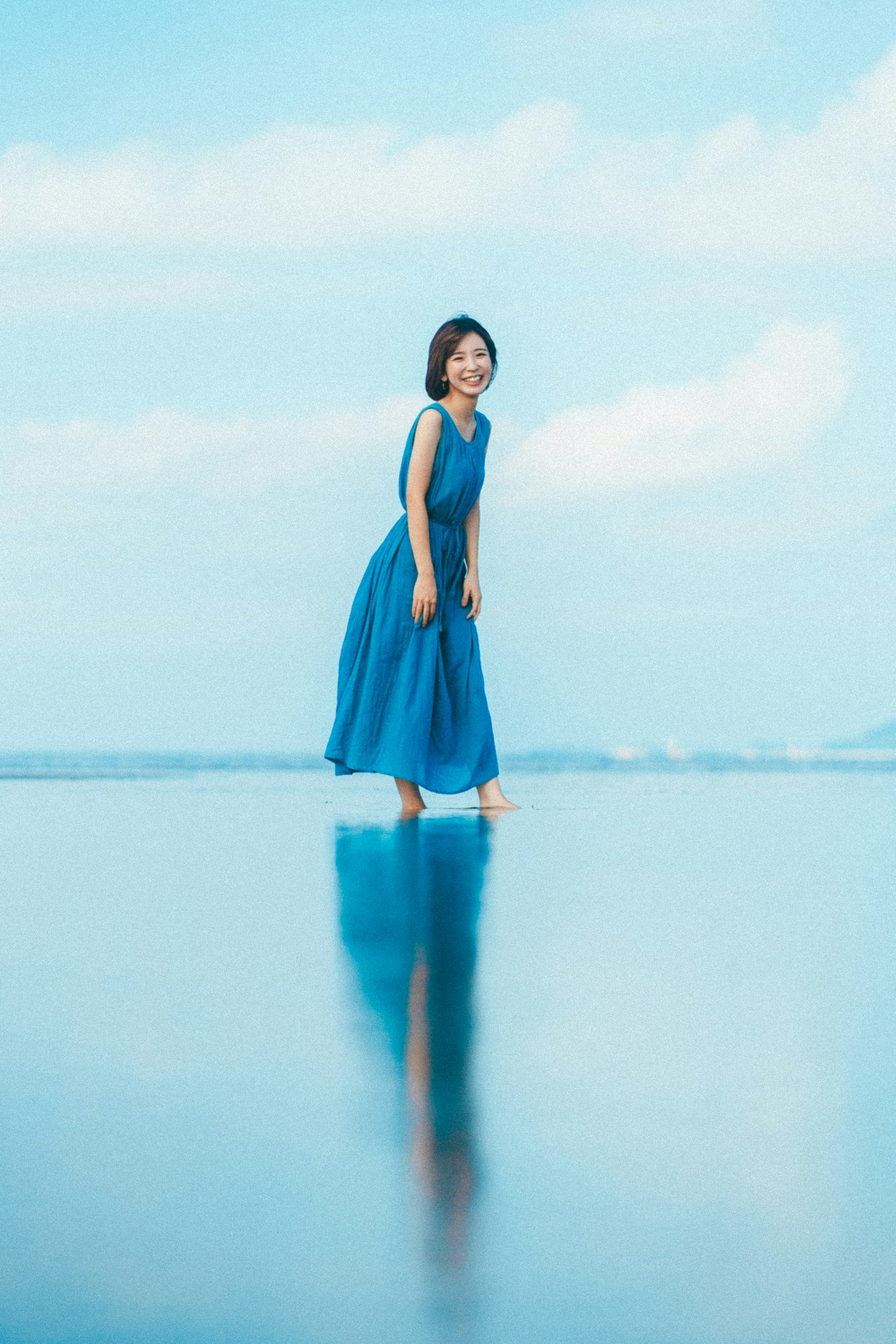 A woman in a blue dress standing on a reflective water surface with a clear sky