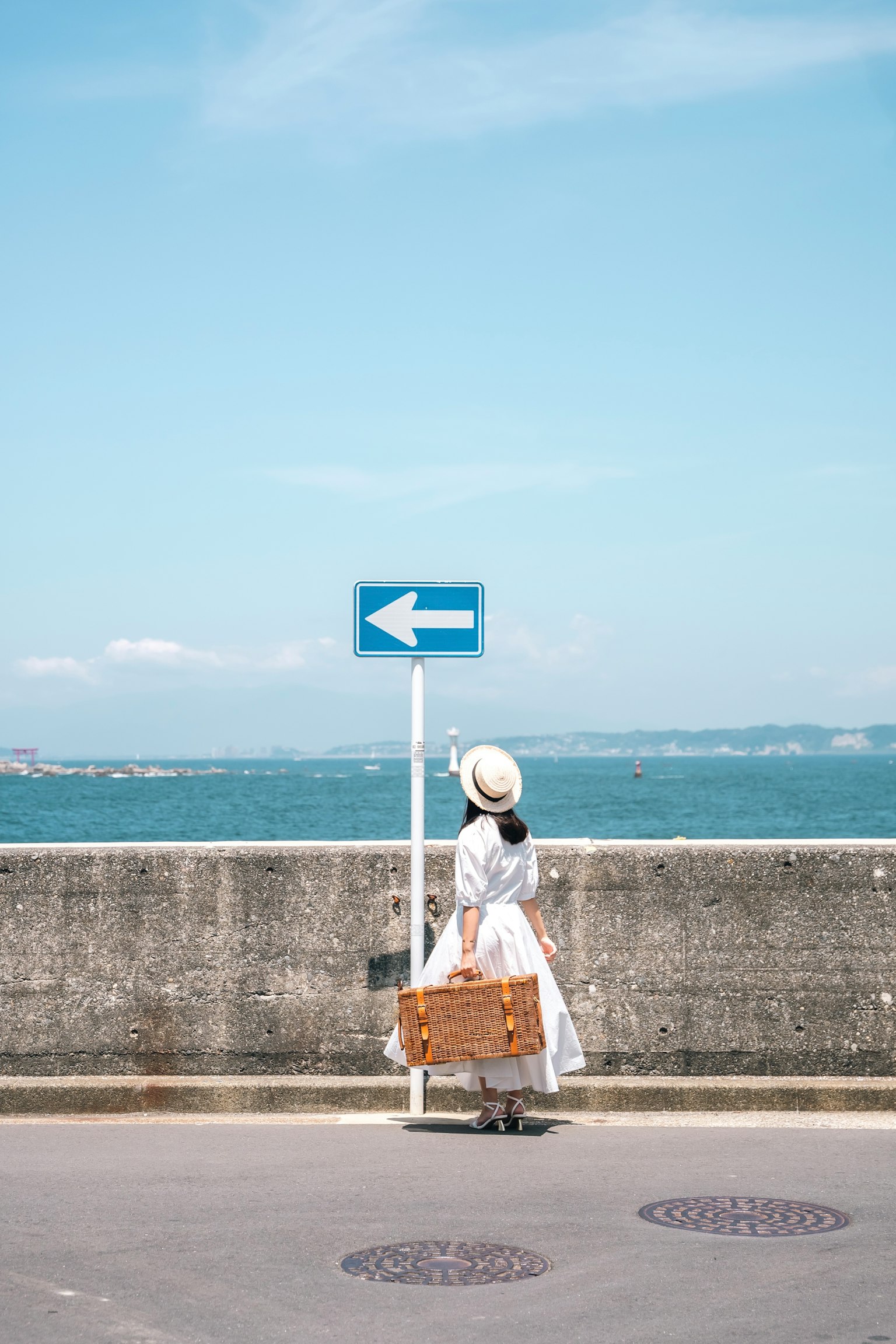 穿著白色裙子的女性站在指向左方的標誌前,背景是海洋