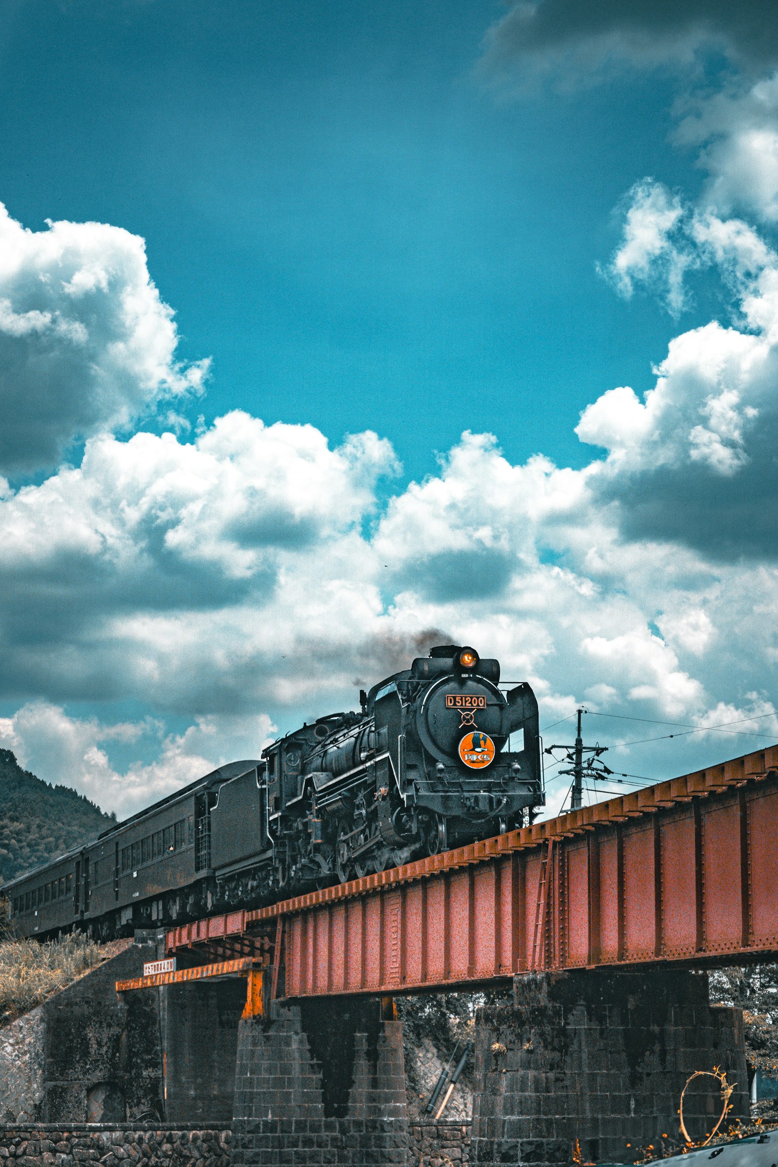 Locomotiva a vapore che attraversa un ponte rosso sotto un cielo blu con nuvole bianche