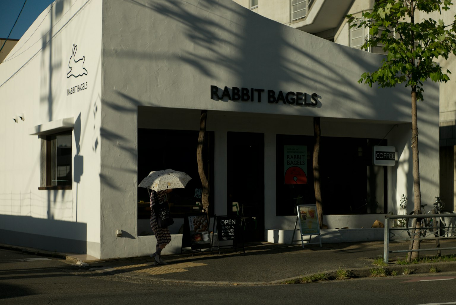Kafe dengan eksterior putih Rabbit Bagels dengan seseorang yang memegang payung di bawah sinar matahari
