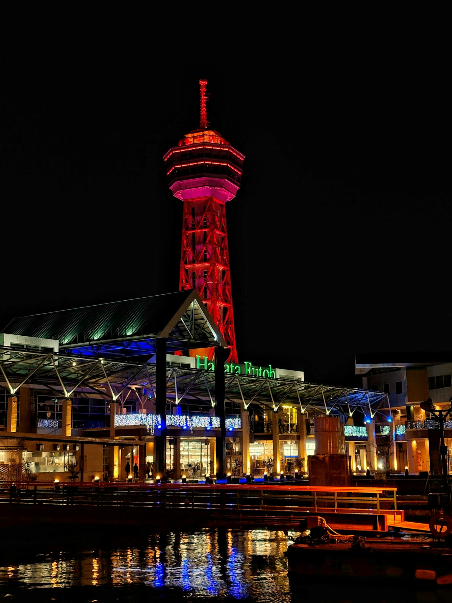 Illuminated tower in red and purple at night with surrounding buildings and reflections in the water