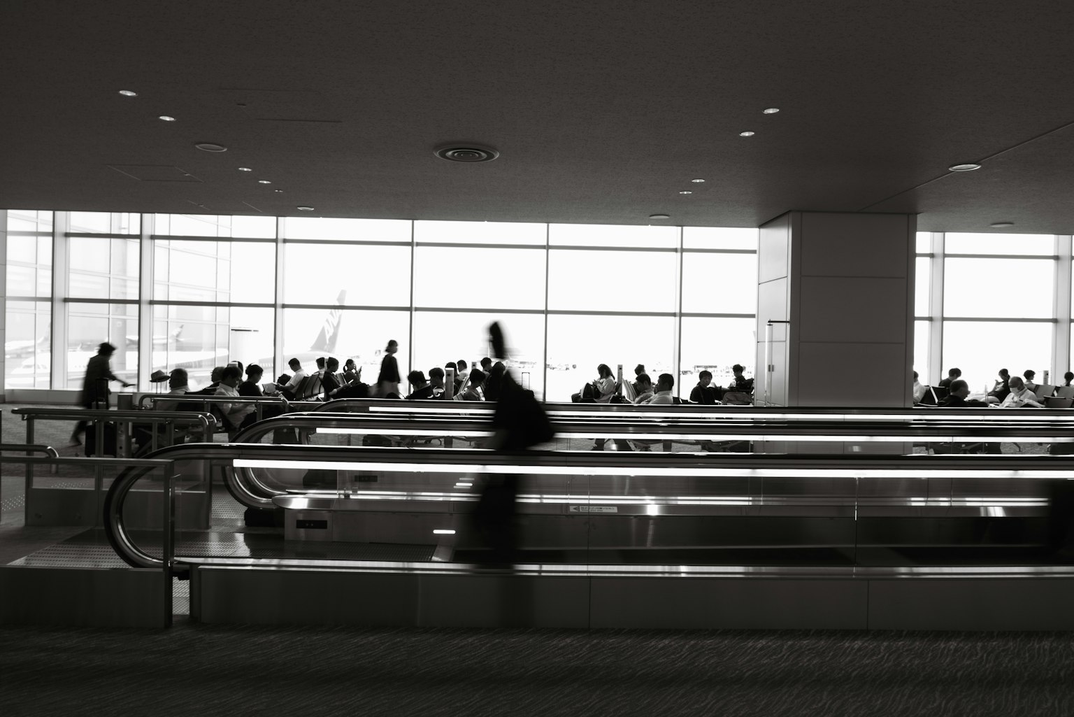 Black and white photo of people in an airport waiting area with moving walkways