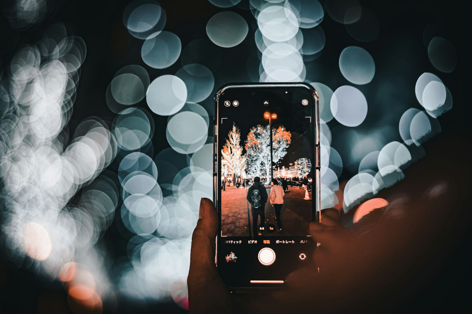 A hand holding a smartphone capturing a photo with blurred lights in the background at night