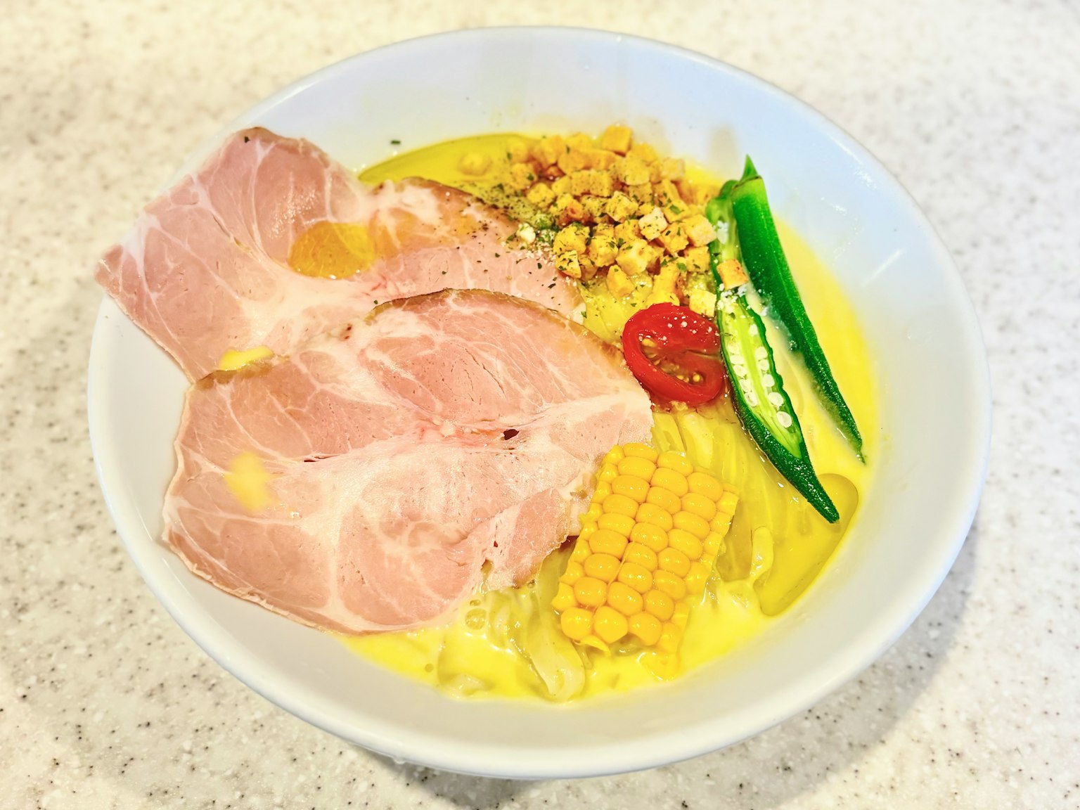 A delicious bowl of ramen topped with sliced pork, corn, okra, tomato, and egg