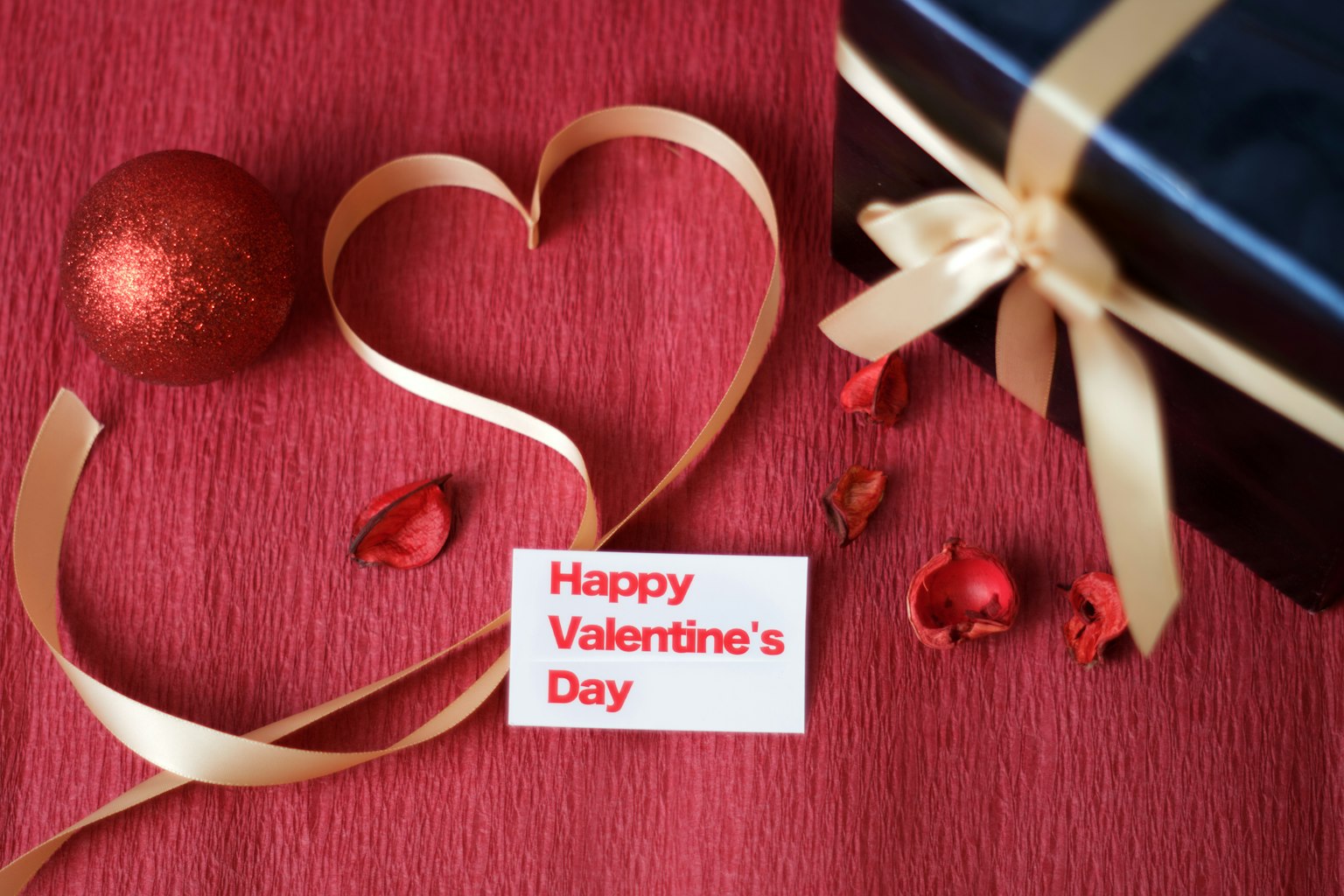Una cinta en forma de corazón con una tarjeta del Día de San Valentín sobre un fondo rojo con una bola roja y pétalos esparcidos