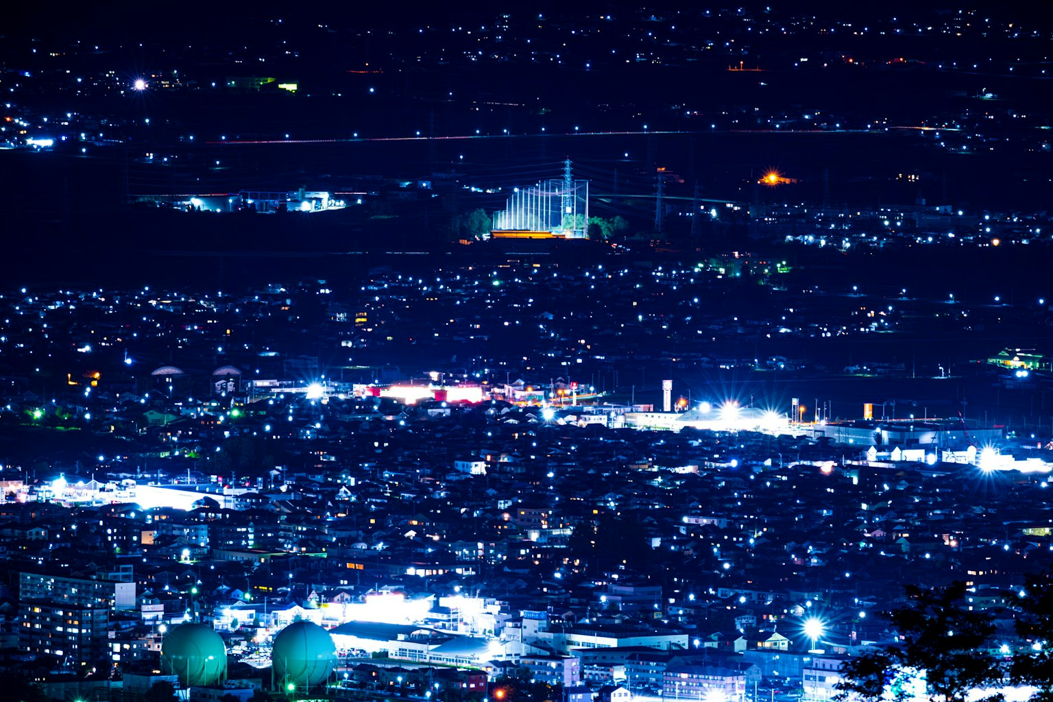 Panorama de la ciudad de noche con edificios iluminados y calles extensas, monumento visible a lo lejos