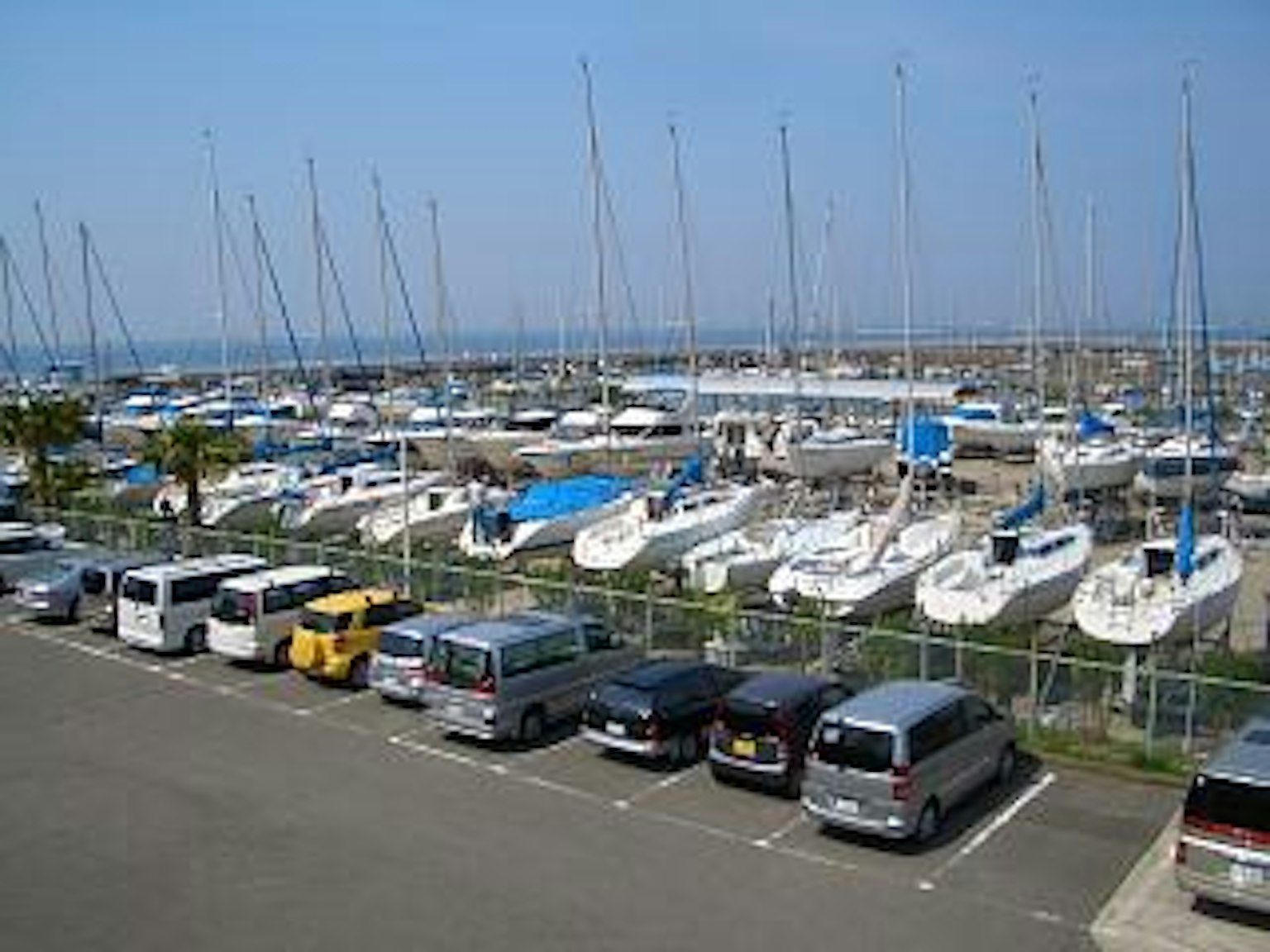 Numerous yachts lined up under a blue sky with parked cars