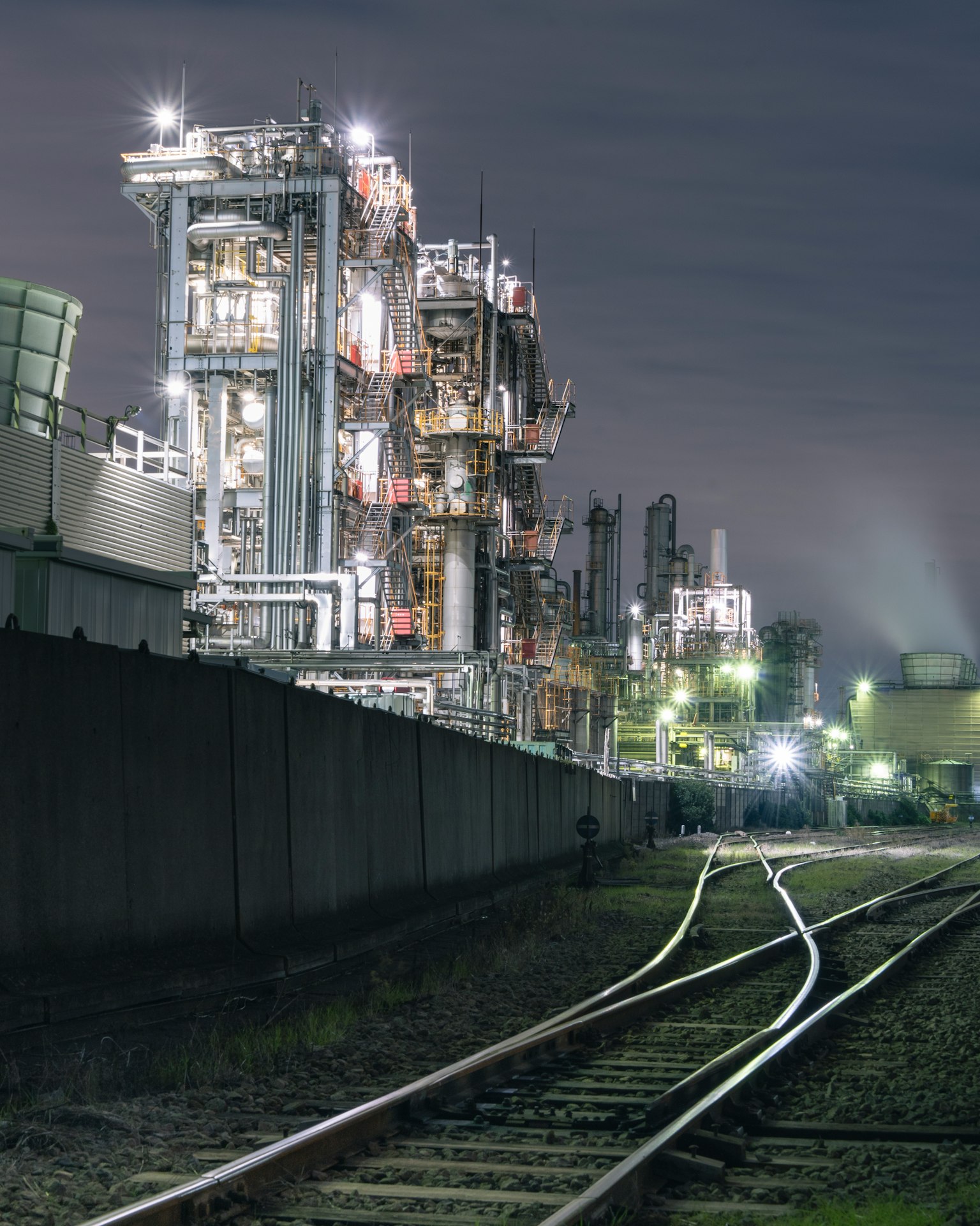 Grande usine industrielle la nuit avec des voies ferrées au premier plan