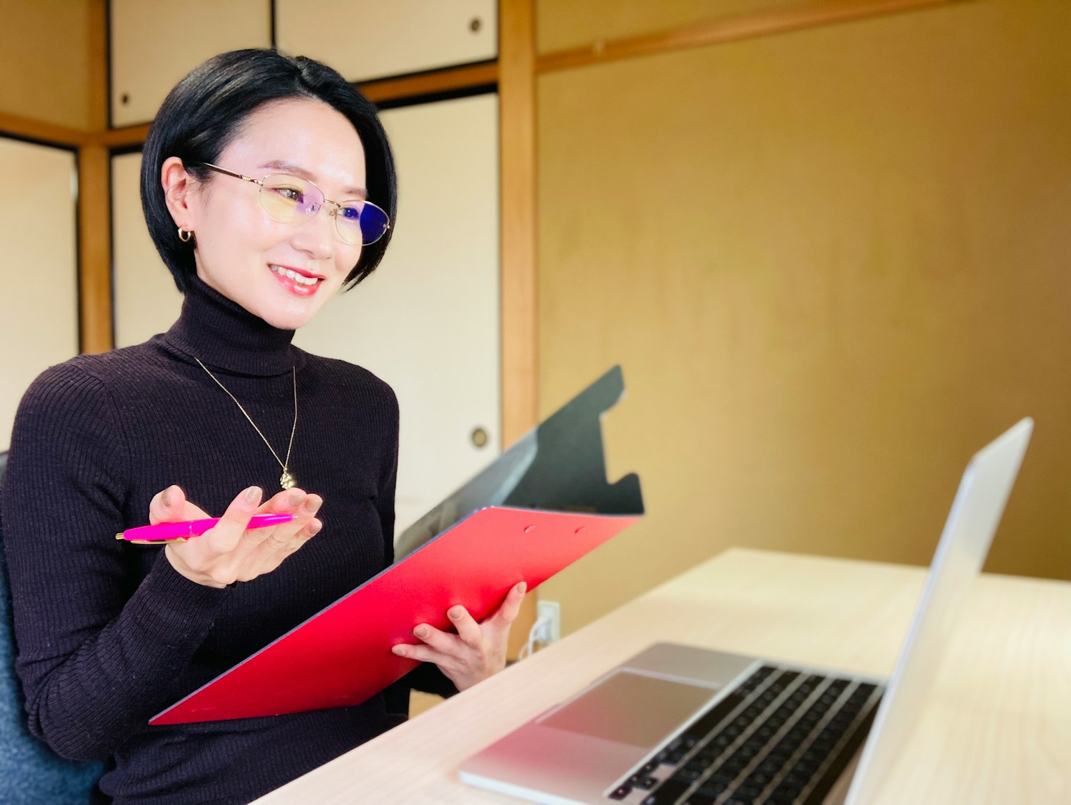 Une femme en col roulé noir tient un clipboard rouge en souriant à un ordinateur portable