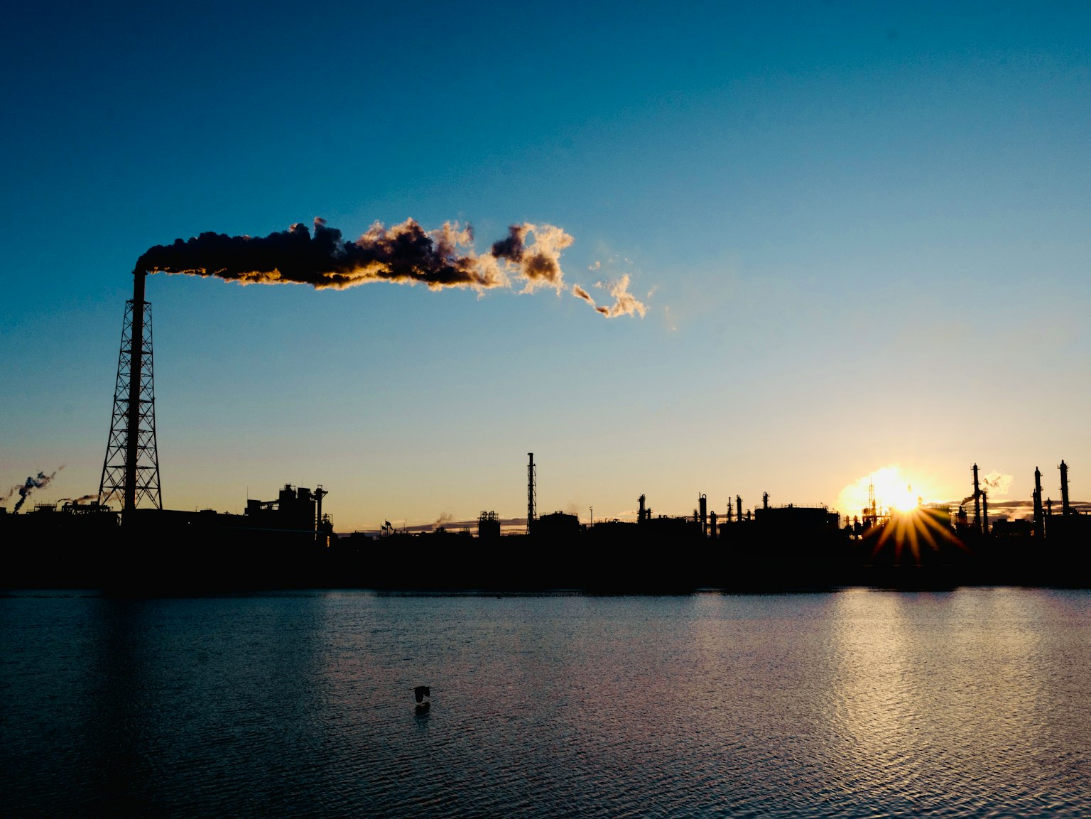 Factory smokestack releasing smoke against a sunset reflected on the water