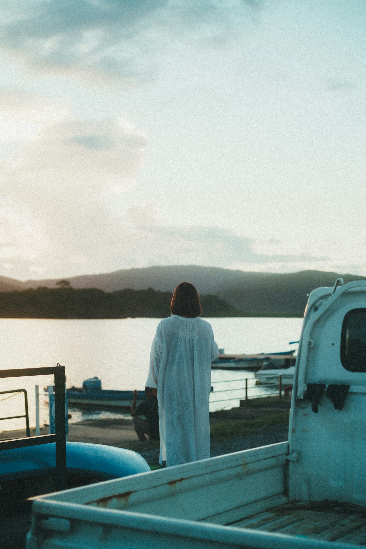 A woman in a white dress stands with her back to the camera by a serene lake at sunset