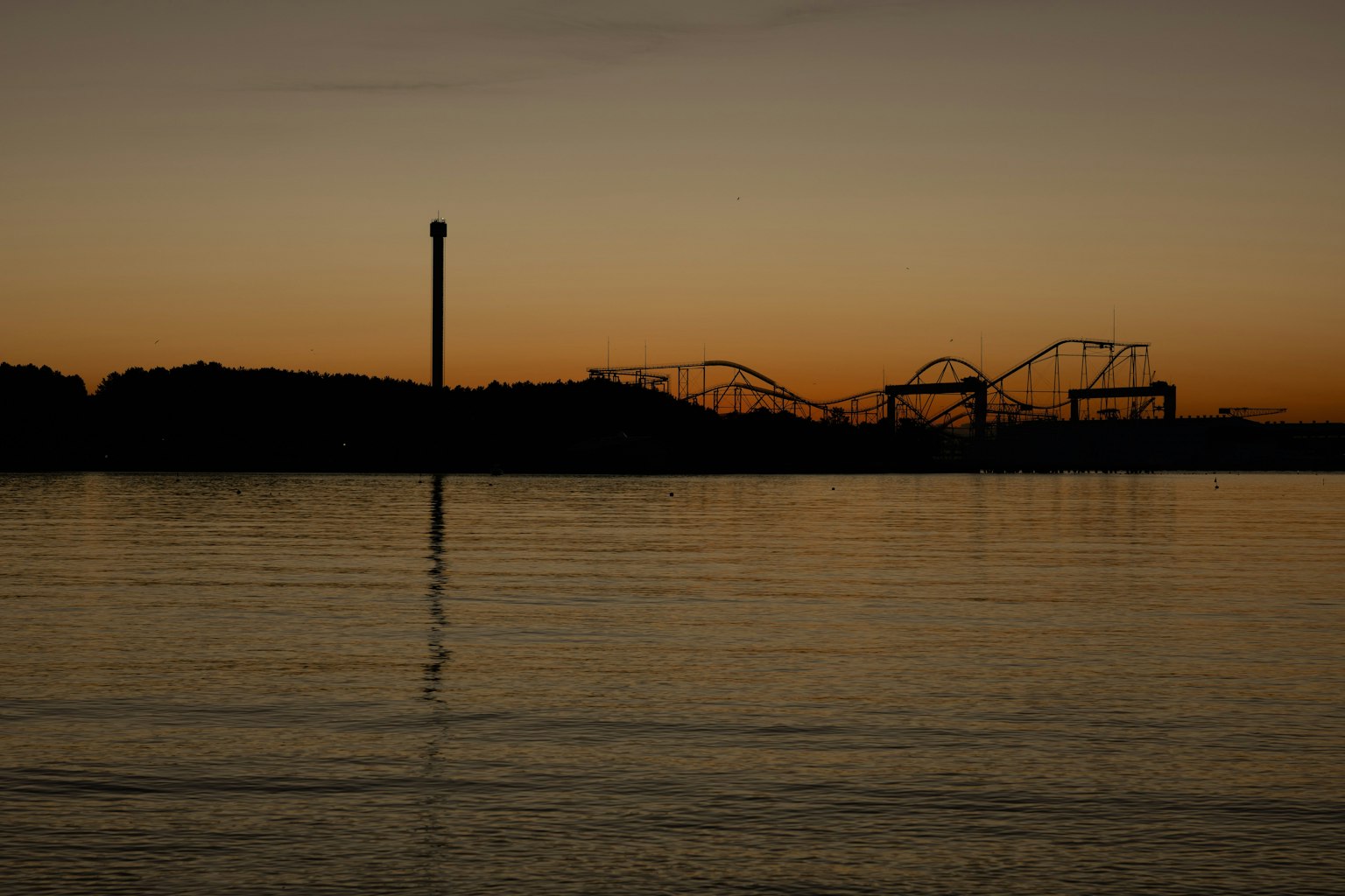 Silhouette of amusement park rides against a sunset with water reflection
