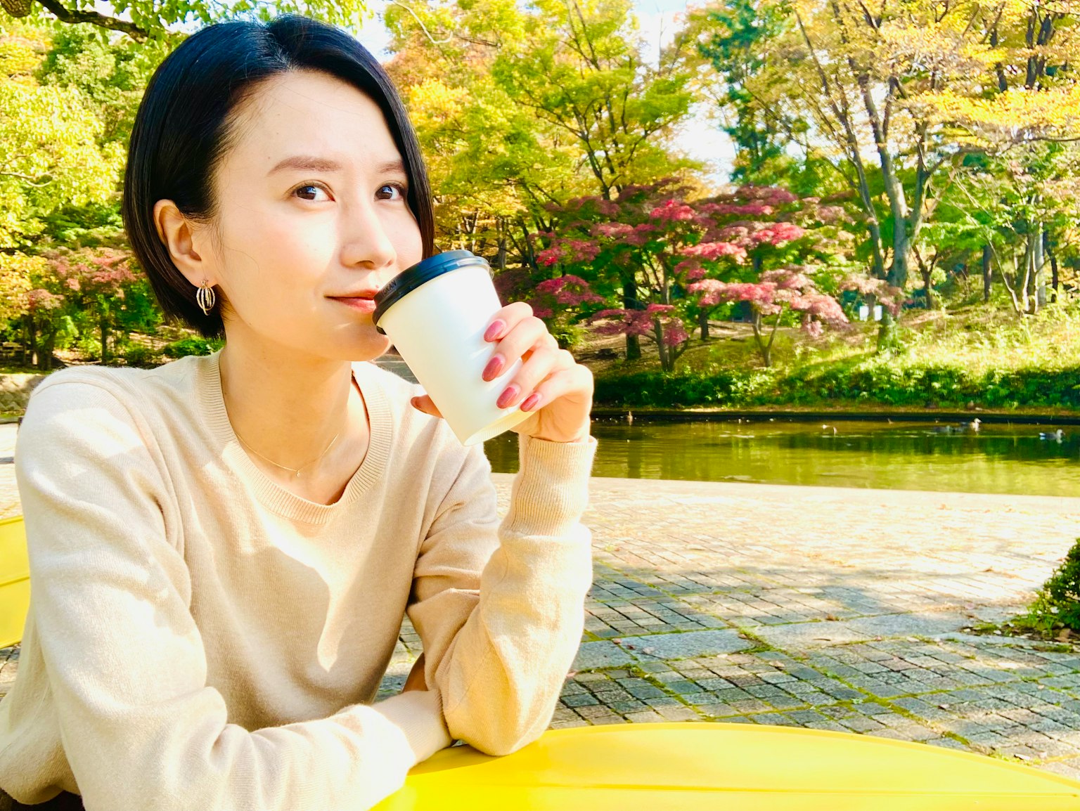 A woman holding coffee in a park with green trees and a pond in the background