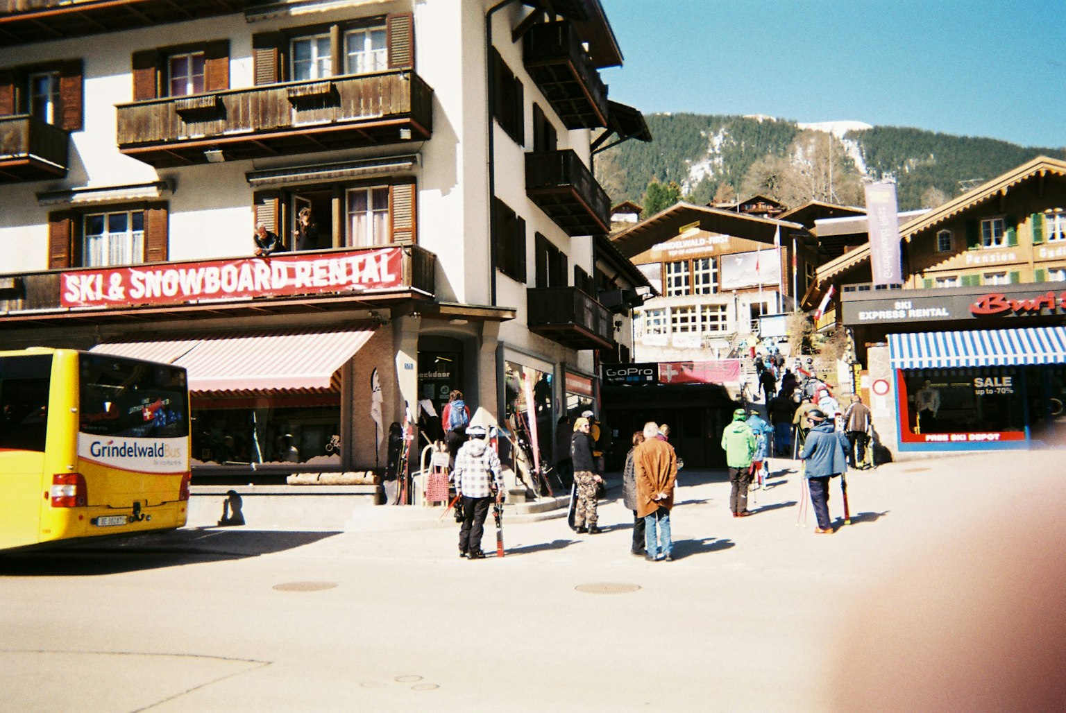Des gens se rassemblent dans une place de village alpin avec un bus jaune et des bâtiments traditionnels