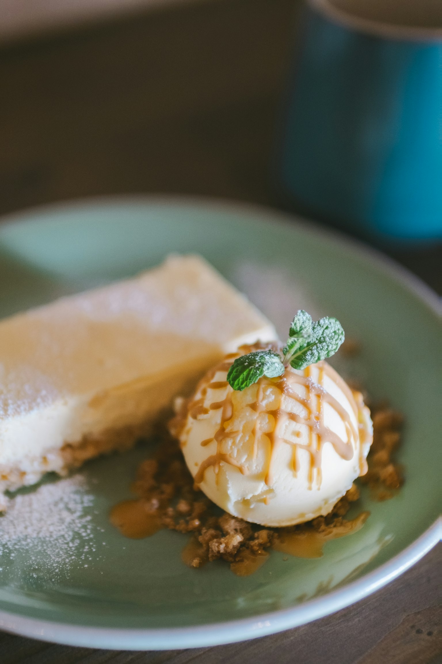 Kue cheesecake krim dengan satu bola es krim vanila disiram saus karamel dan dihias dengan daun mint