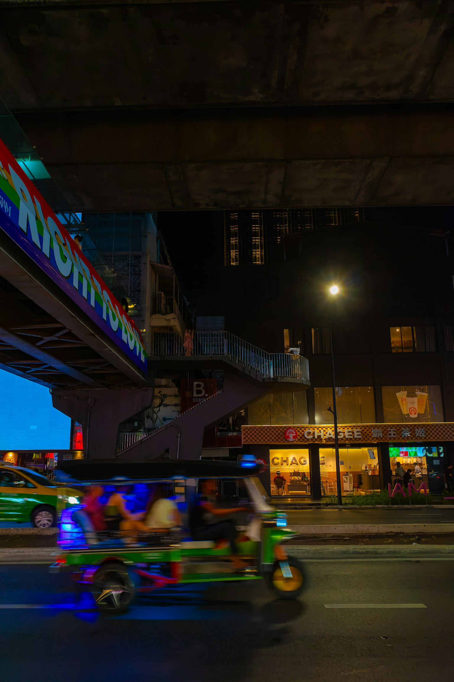夜晚的街道上，三轮车在桥下行驶，周围有五彩缤纷的招牌和商店灯光