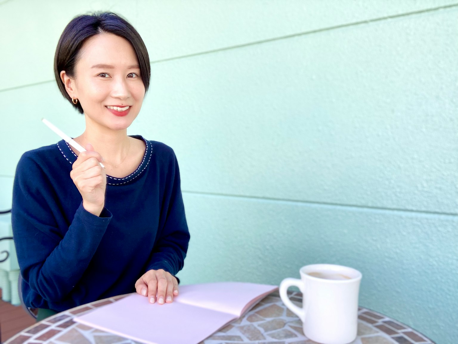 Một người phụ nữ trong áo len màu xanh đang mỉm cười trong khi cầm sổ tay và bút ở bàn