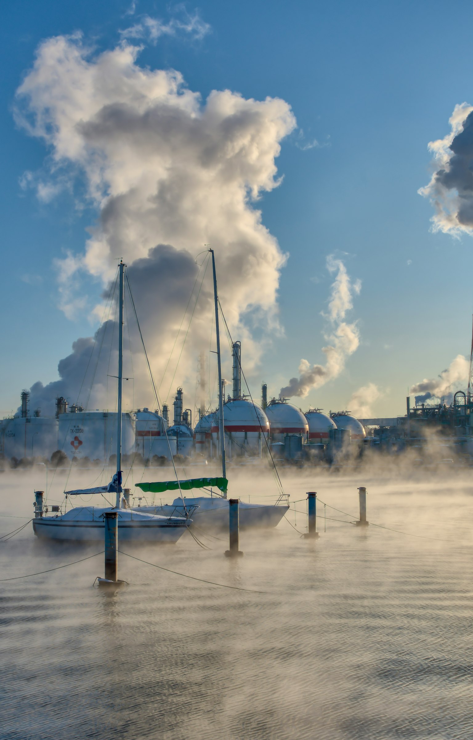 A yacht emerging from mist with smoke billowing from industrial buildings