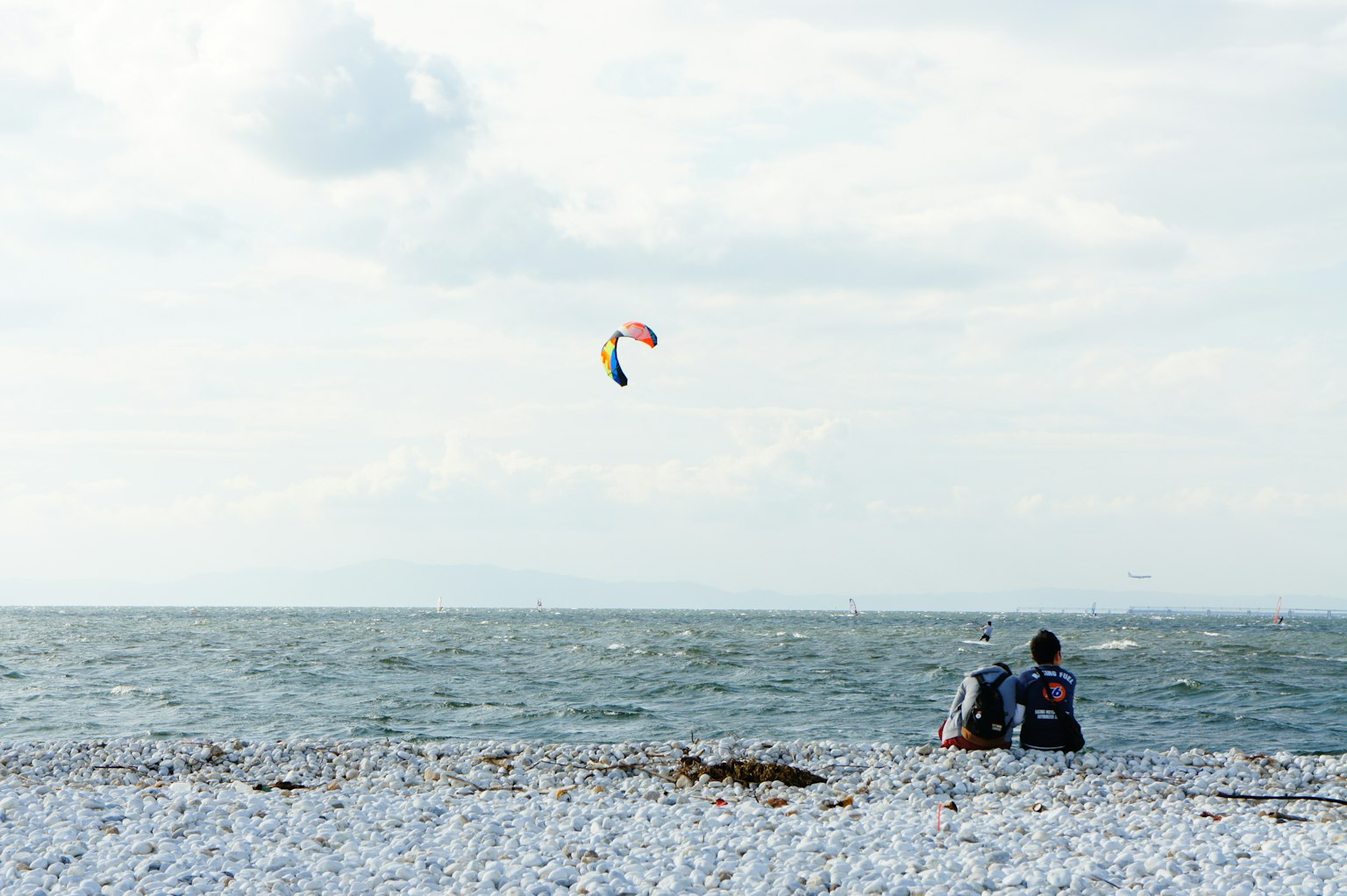 Strandszene mit einer Person, die auf Kies sitzt, und einem bunten Drachen am Himmel