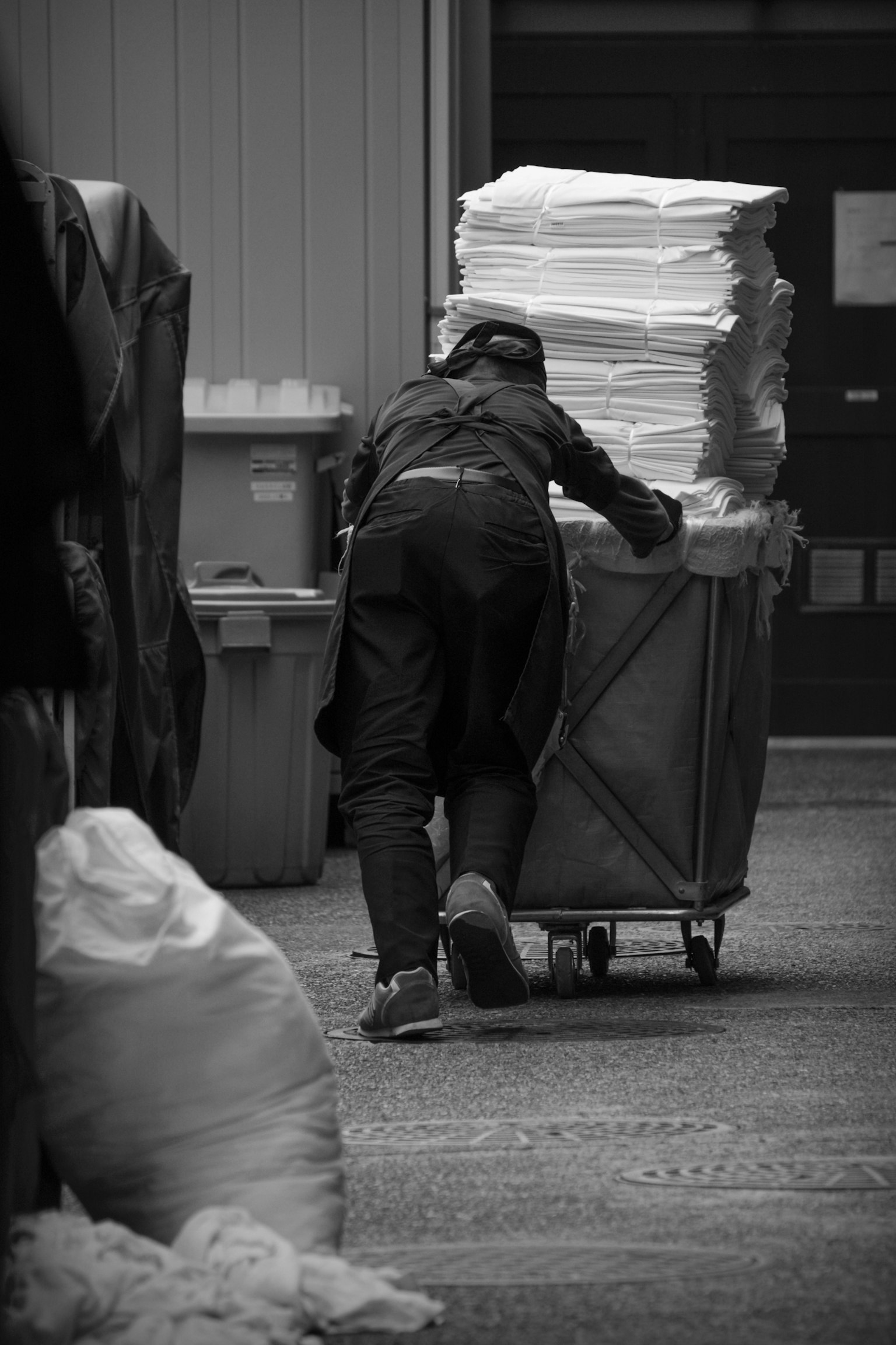 A black and white image of a worker pushing a cart filled with sheets