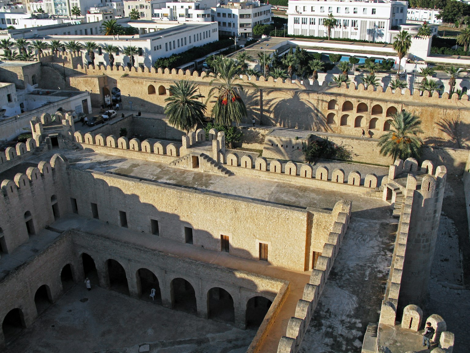 Vue aérienne d'une ancienne forteresse avec des murs en pierre et des tours entourées de palmiers et de bâtiments modernes