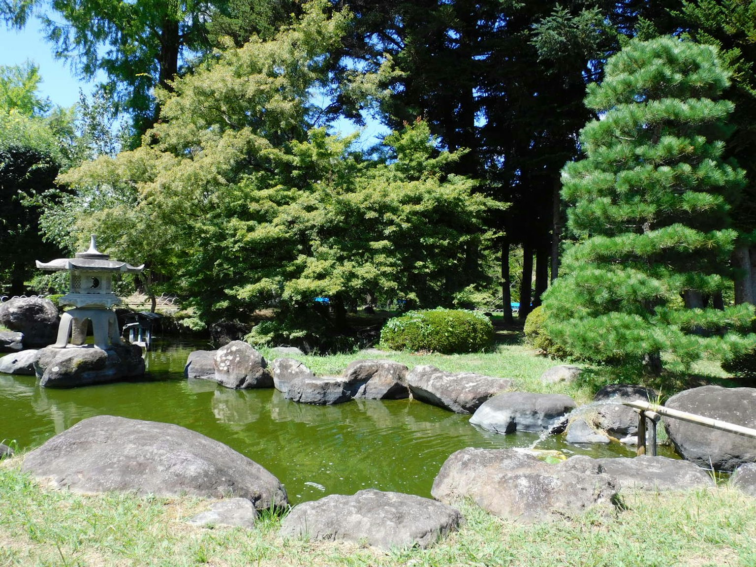 Taman Jepang dengan kolam dan lentera batu dikelilingi oleh tanaman hijau subur dan batu