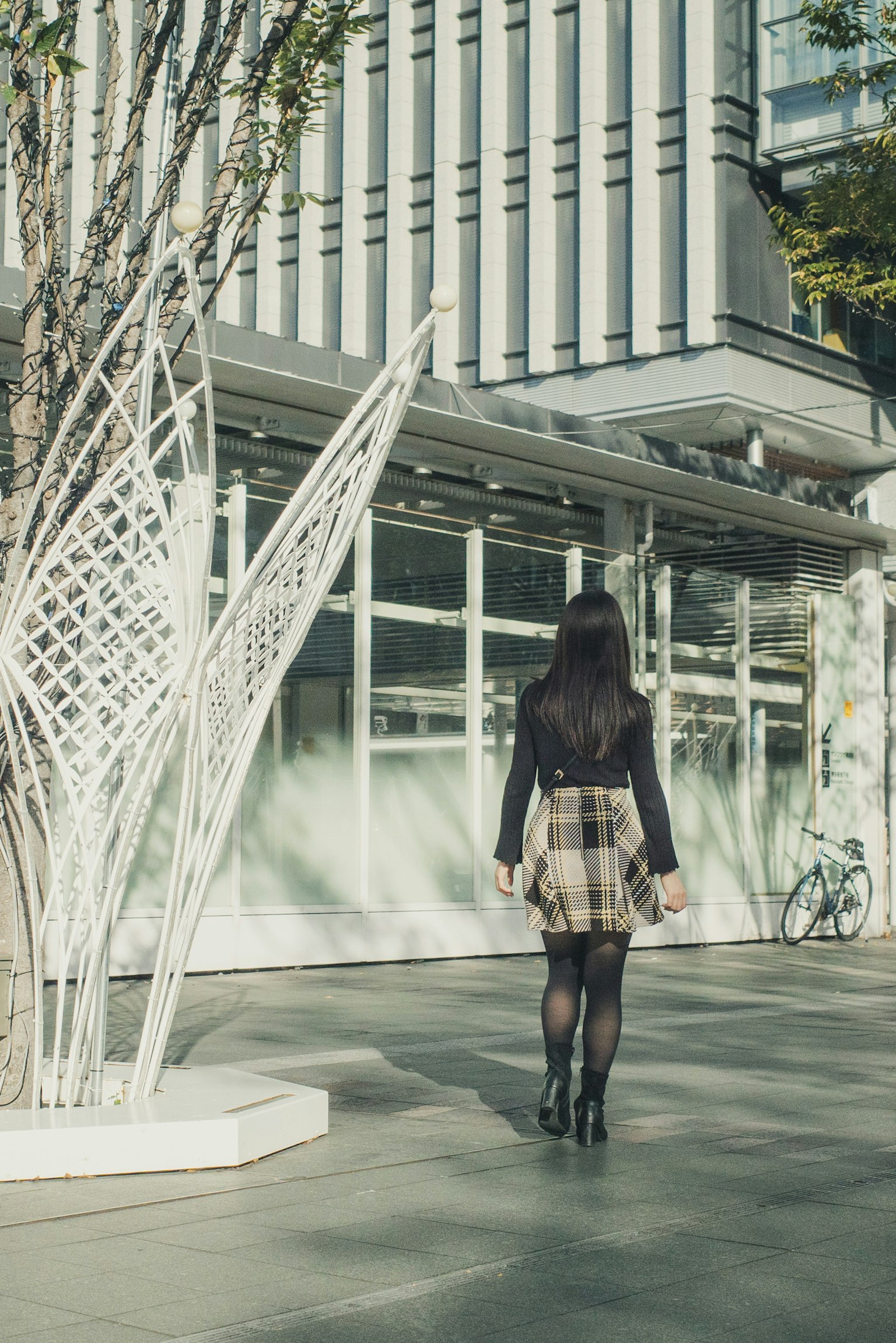 A woman in a checkered skirt stands with her back to a modern building