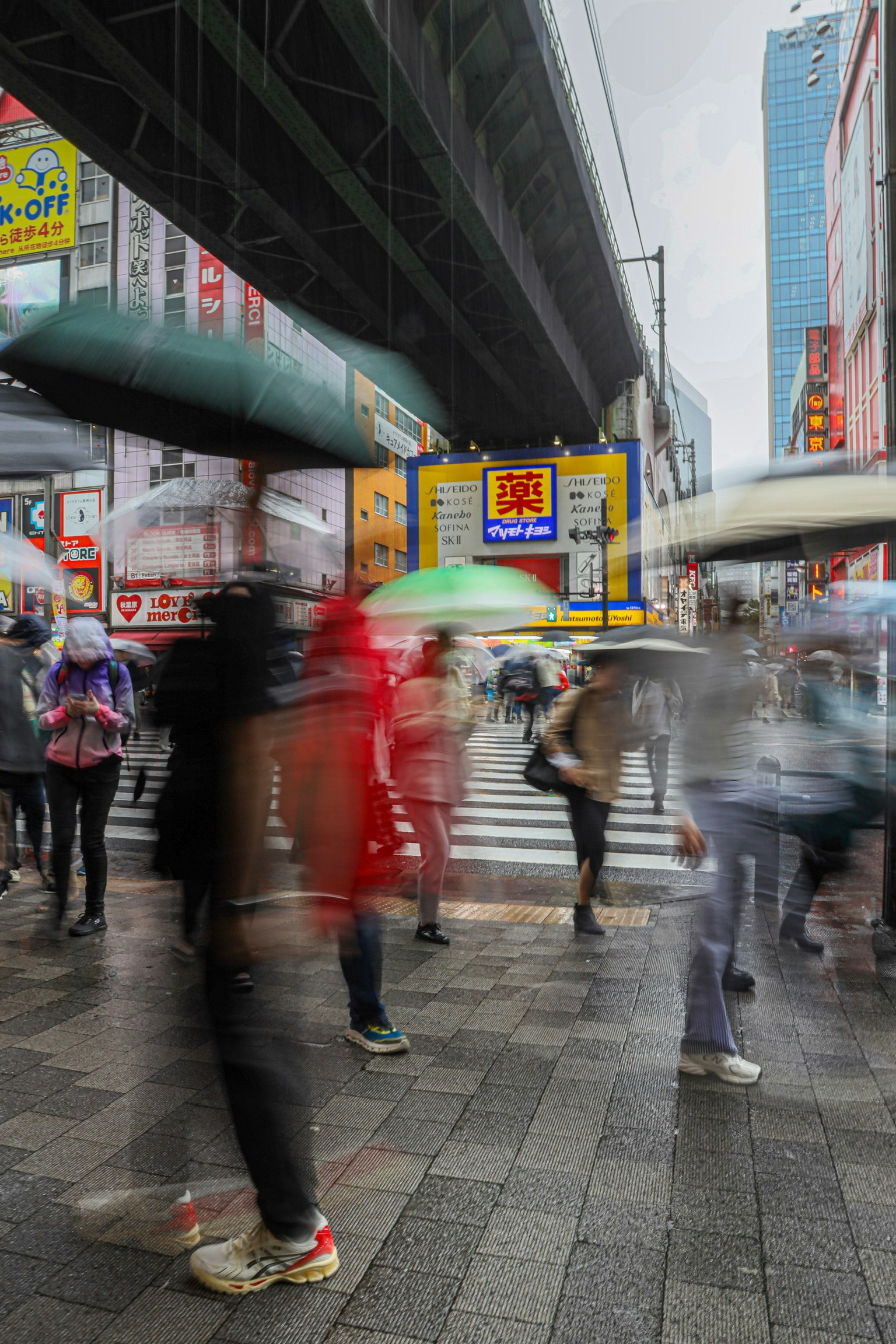 雨の中で傘を持った人々が交差点を渡る様子 近くのビルや広告が見える