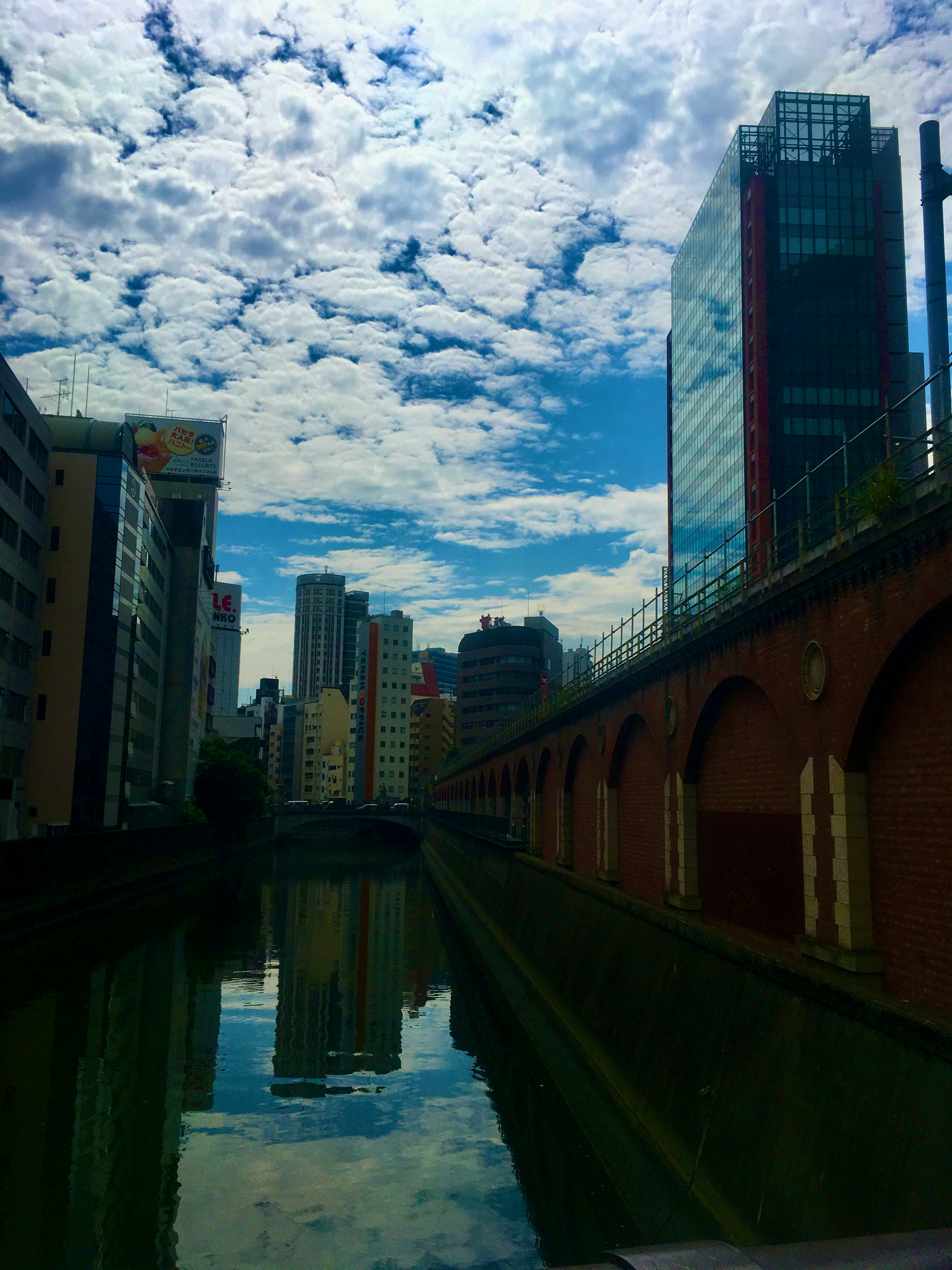 ビルの間に広がる雲と青空、川沿いの水面