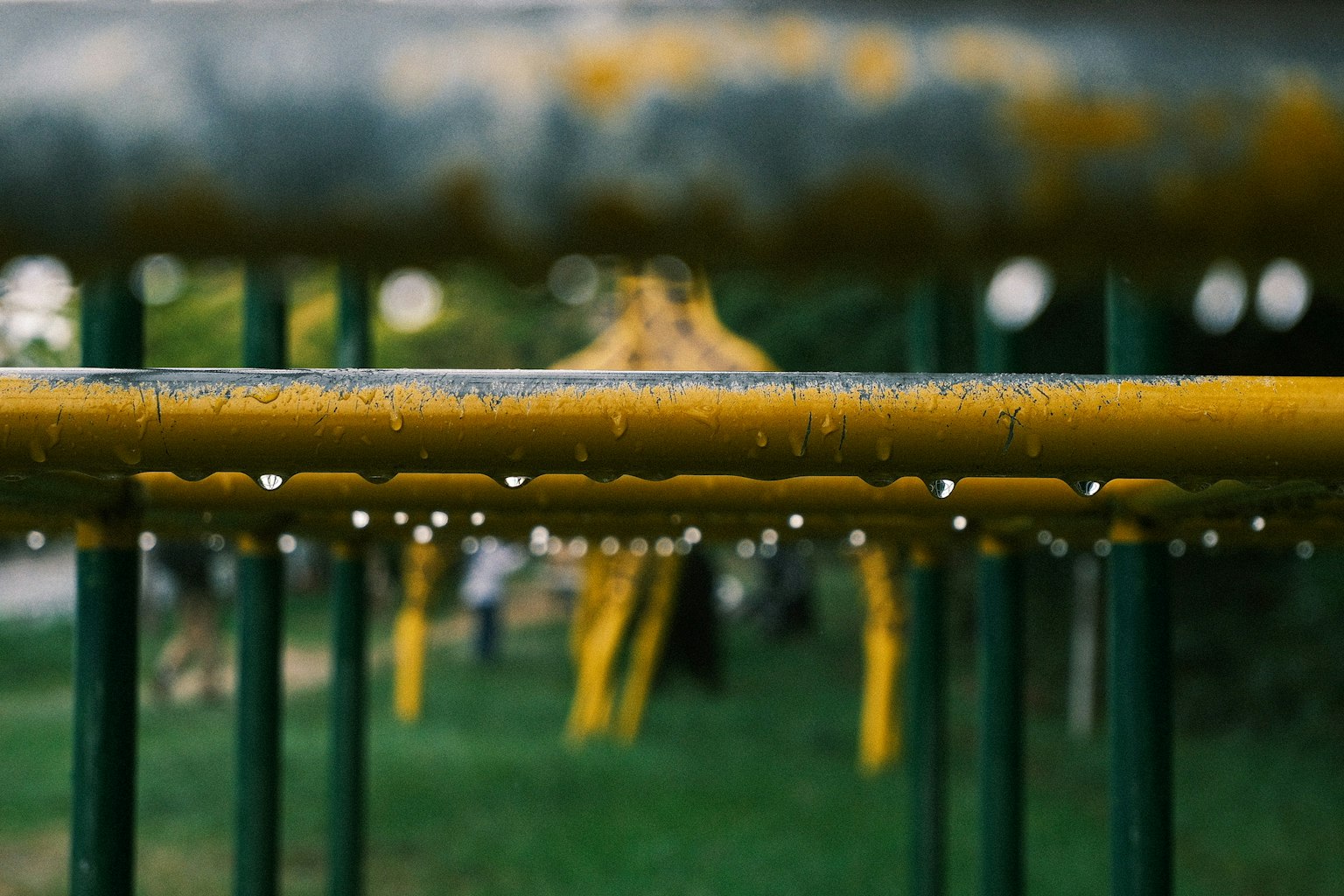 公園の遊具の黄色いバーに水滴が付いている写真 背景には緑の草とぼんやりした人々が見える