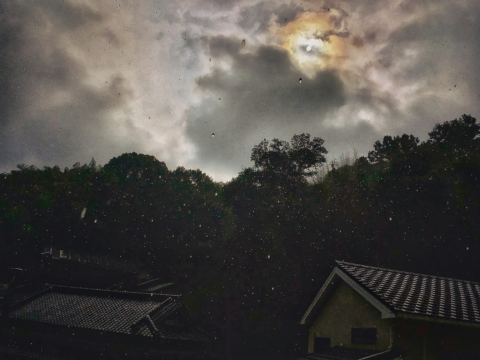 Ein dunkler Himmel mit Wolken und fallendem Regen über Häusern mit einem schwachen Mond sichtbar